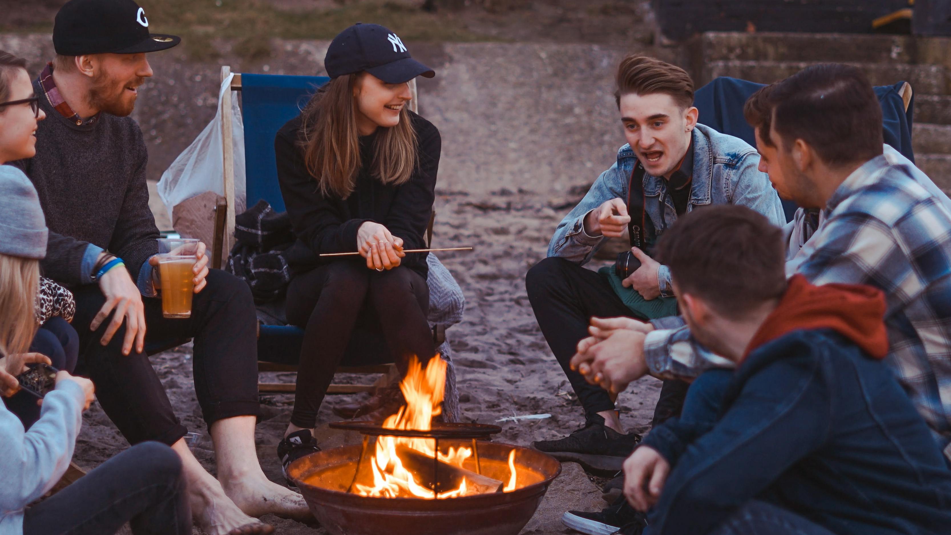 Several people sit around a campfire in camp chairs together. 