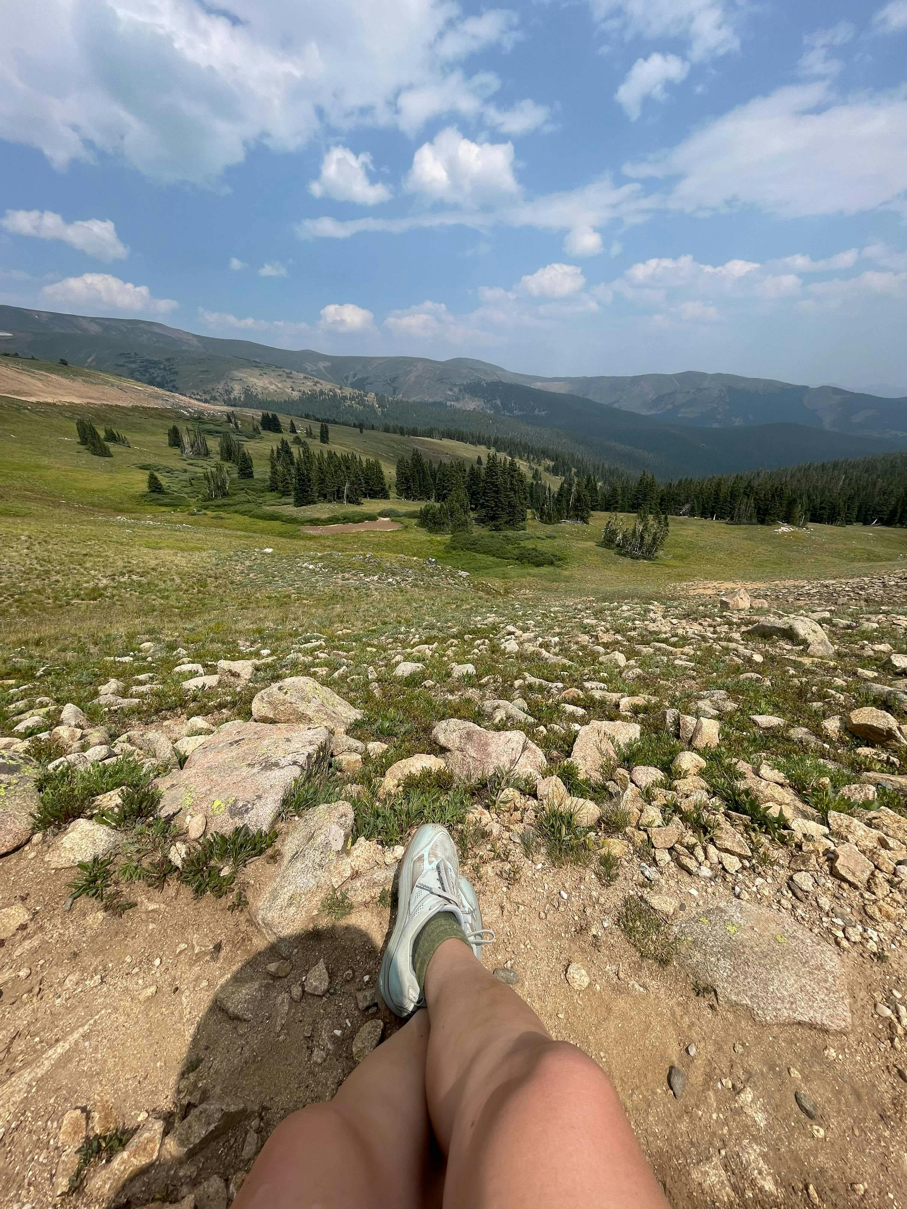 The author sits and we see her legs outstretched before an expansive view of the trail, a meadow, and hills in the distance. The sky is blue with white fluffy clouds. 