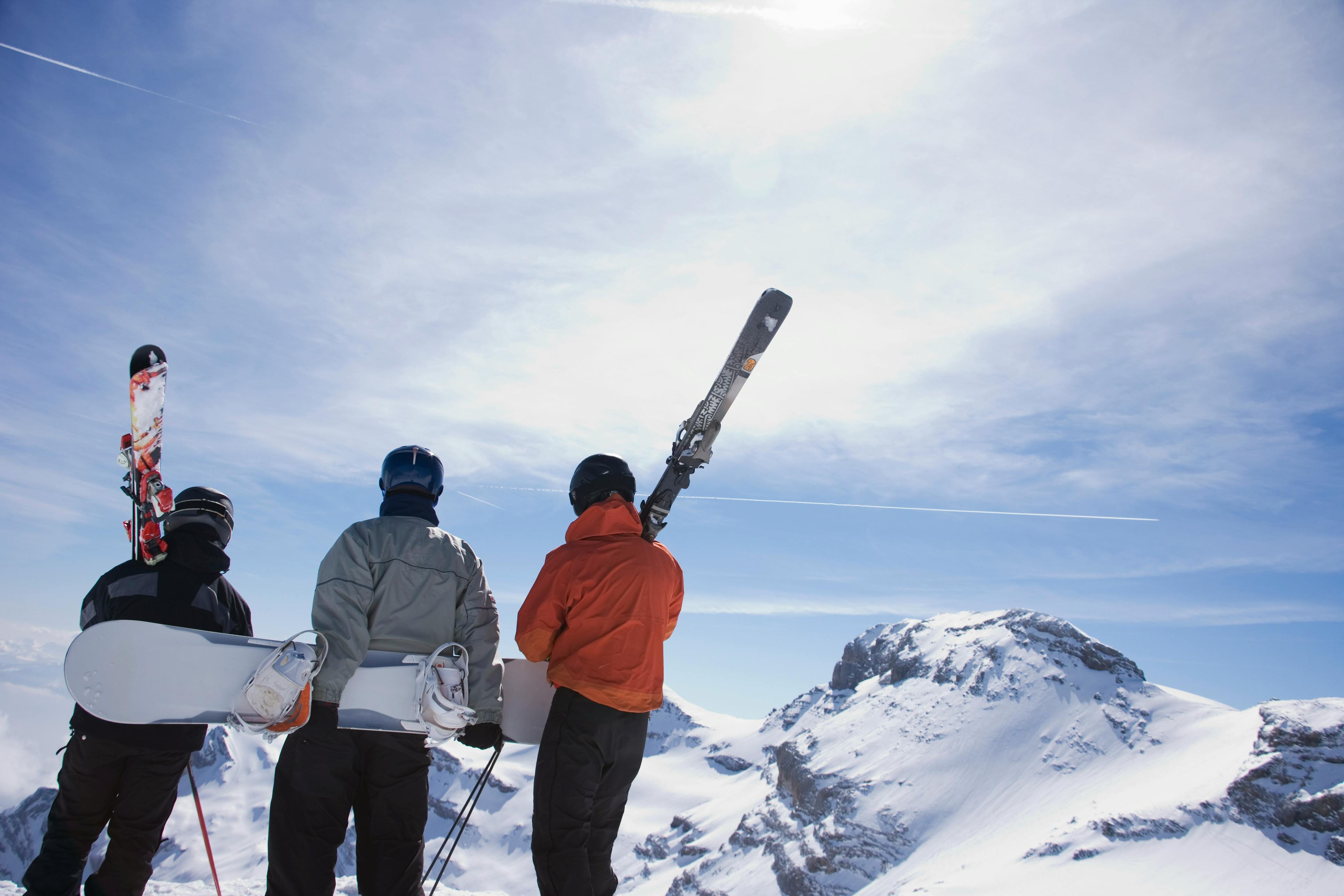 Two skiers and a snowboarder stand on the edge of a hill looking out at snowy mountains