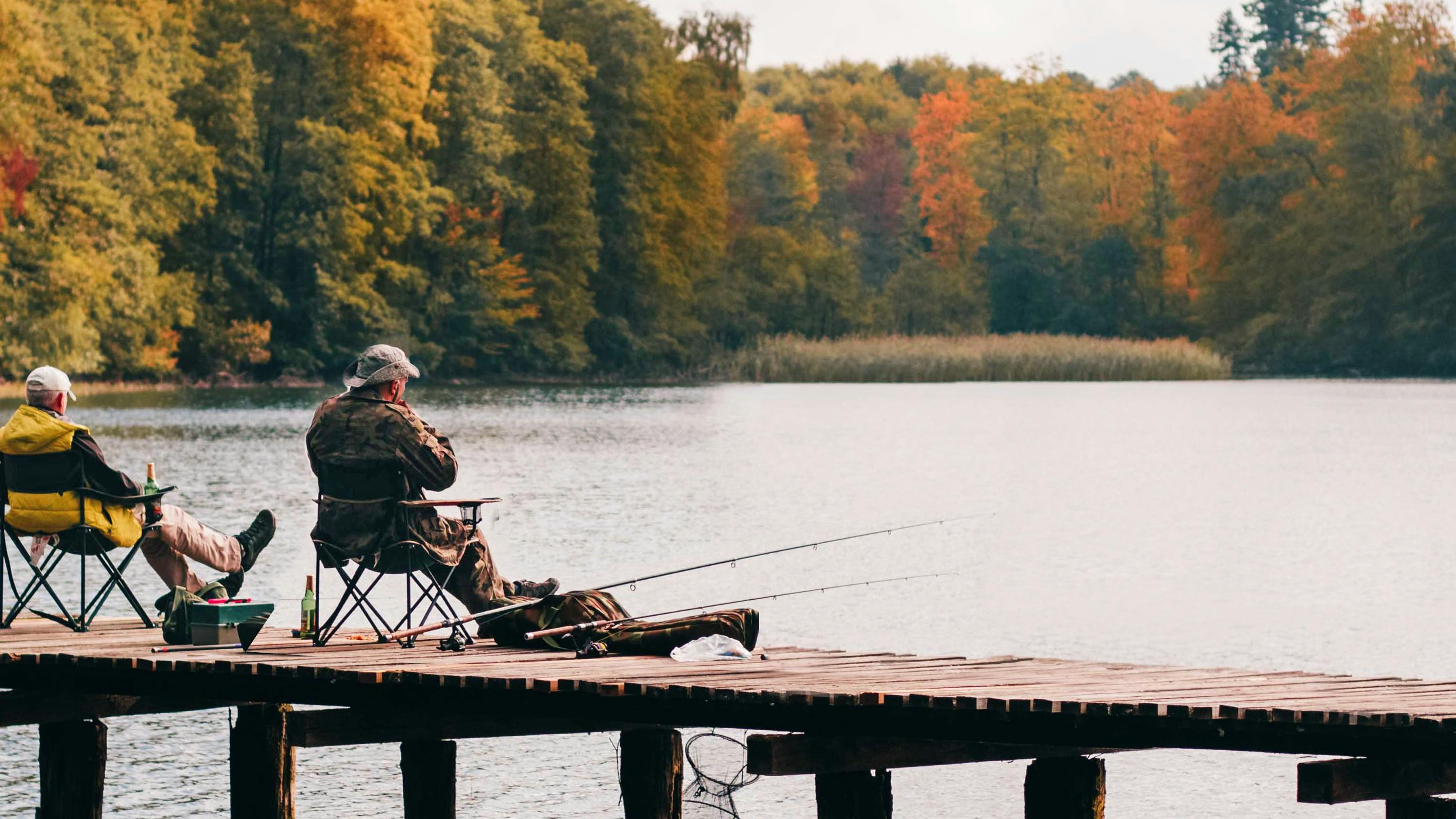 Two men sit on a dock and fish. The trees surrounding the pond are changing colors.