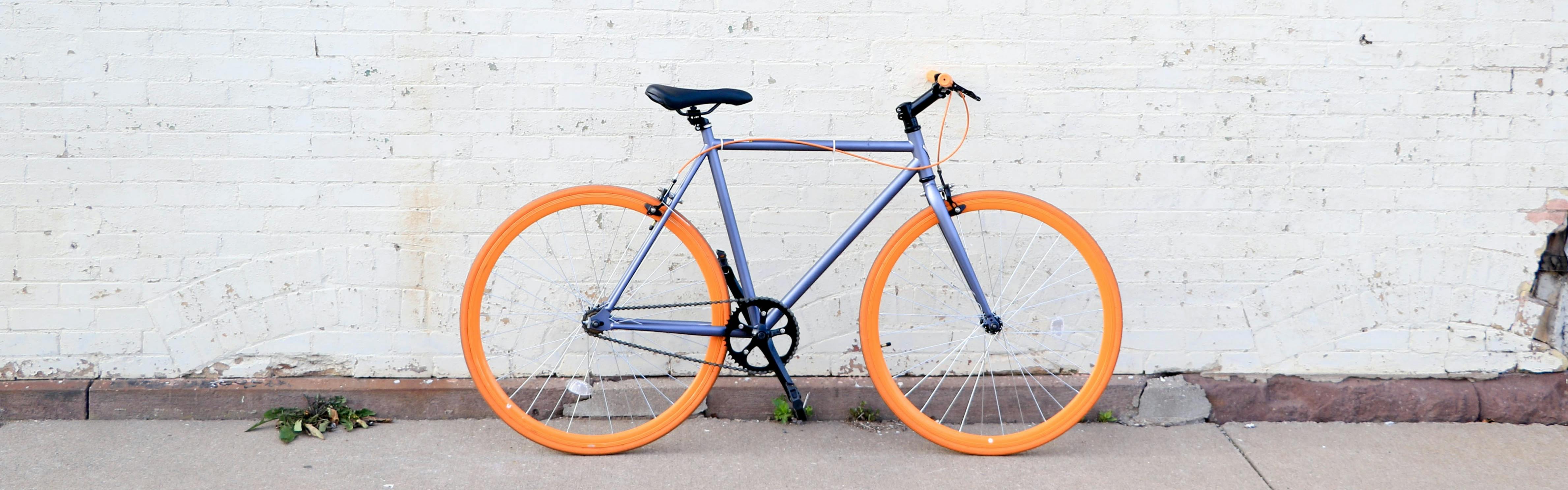A purple bike with vibrant orange tires standing on the sidewalk against a blank white wall.