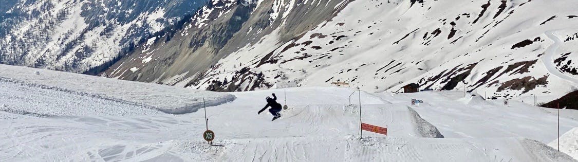 A woman on a snowboard goes off a small jump on a ski run. There are snowy mountains in the background.