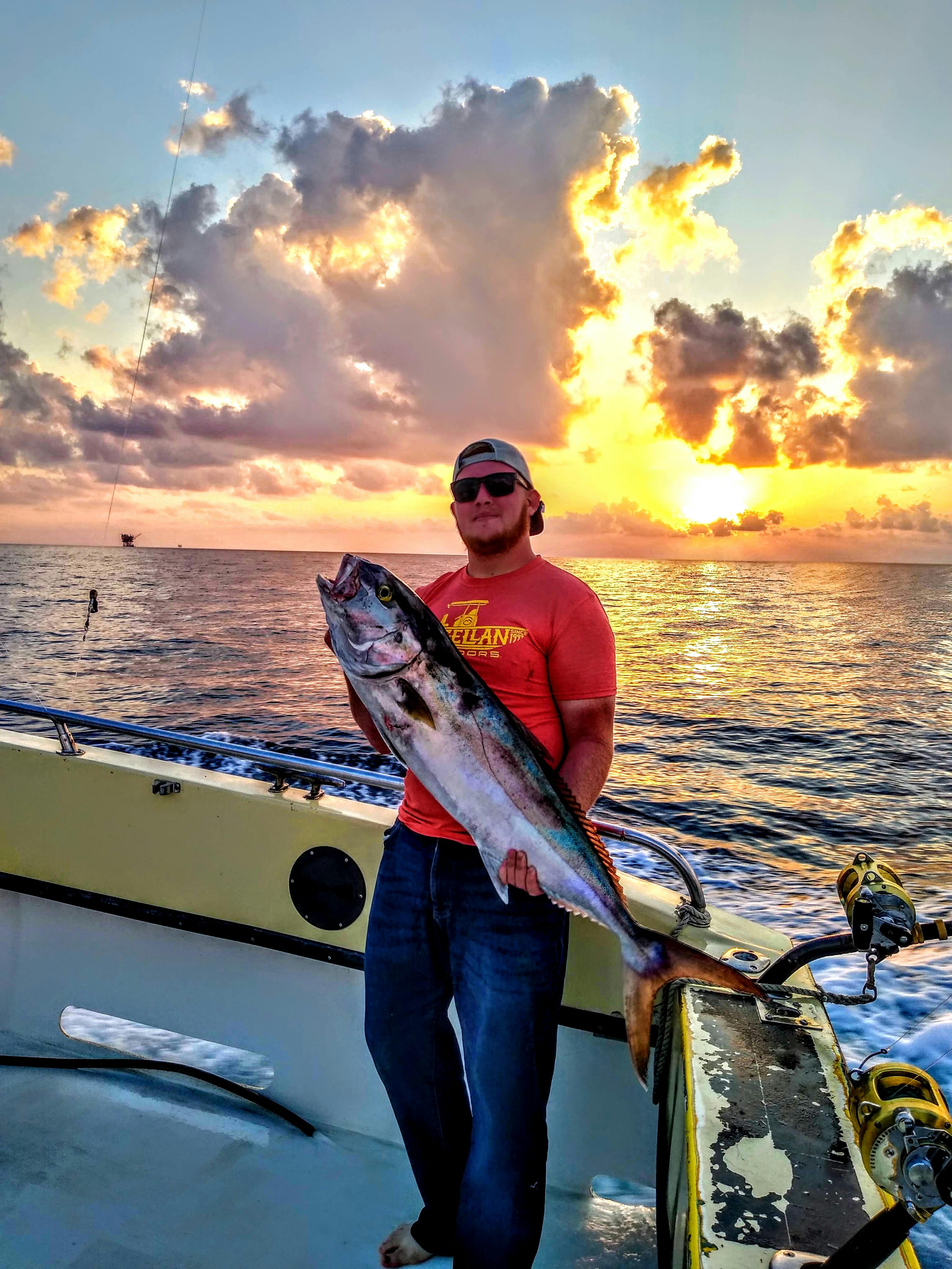 The author stands on a boat on open water, holding a very large fish and posing in front of a brilliant sunset. 