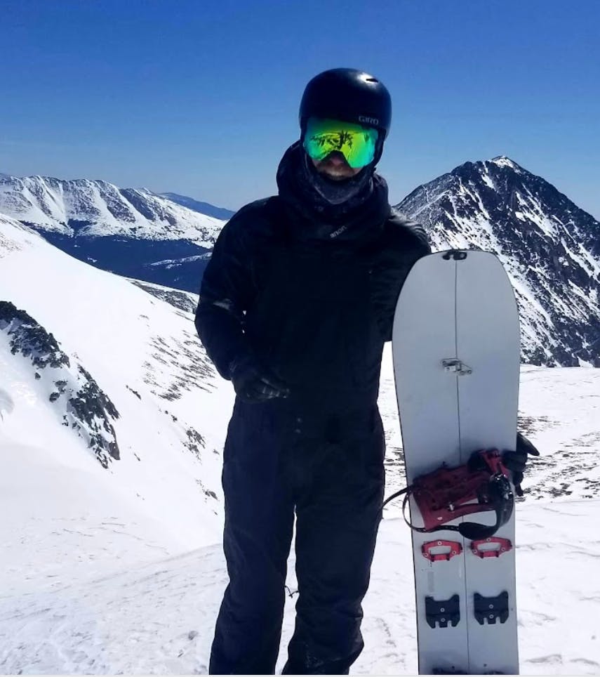 A man standing at the top of a peak with his snowboard and gloved hands. 