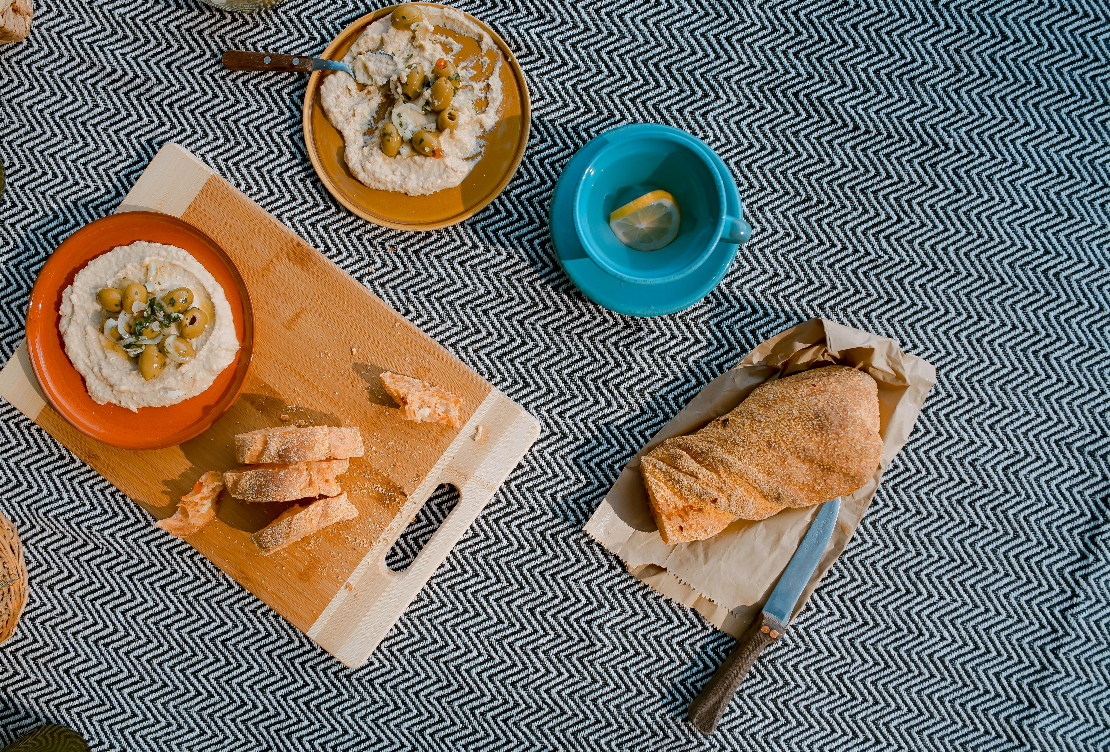 Hummus and bread on a cutting board, which is laying on a picnic blanket. 