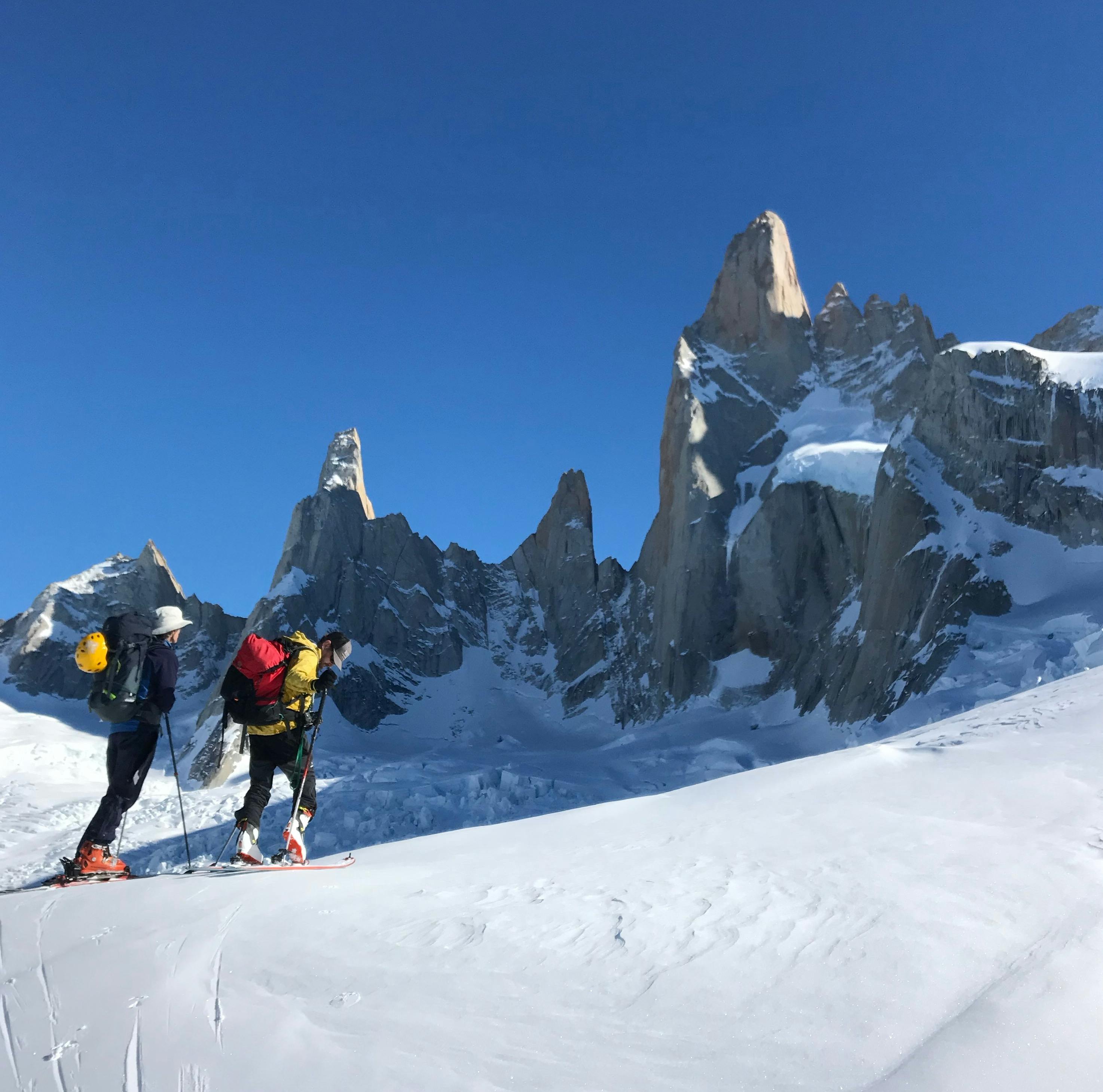 Two people ski below jagged peaks.
