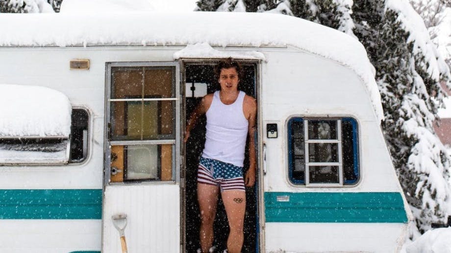 Mogul skiing Olympian Casey Andringa stands in the doorway of the camper that he lived in while preparing for the Olympics. The camper is covered in snow and he is wearing American flag shorts.