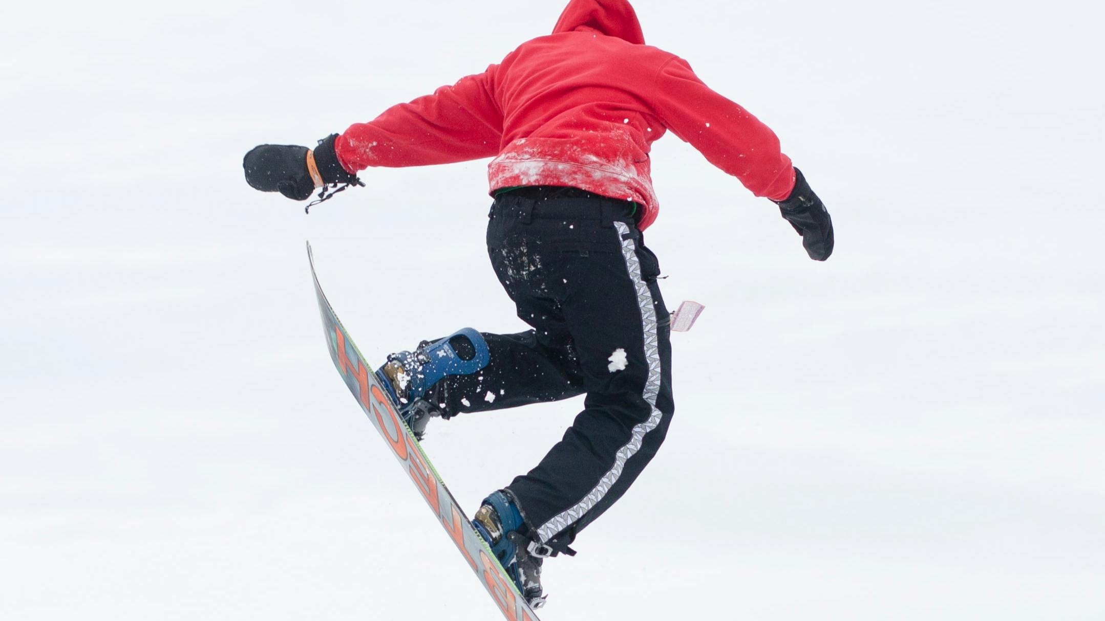 Snowboarder in a bright red jacket and black snow pants flying through the air in a snowboard trick.