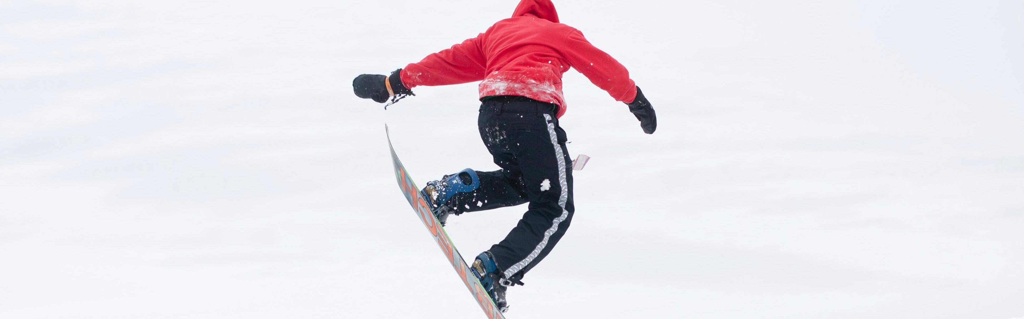 Snowboarder in a bright red jacket and black snow pants flying through the air in a snowboard trick.