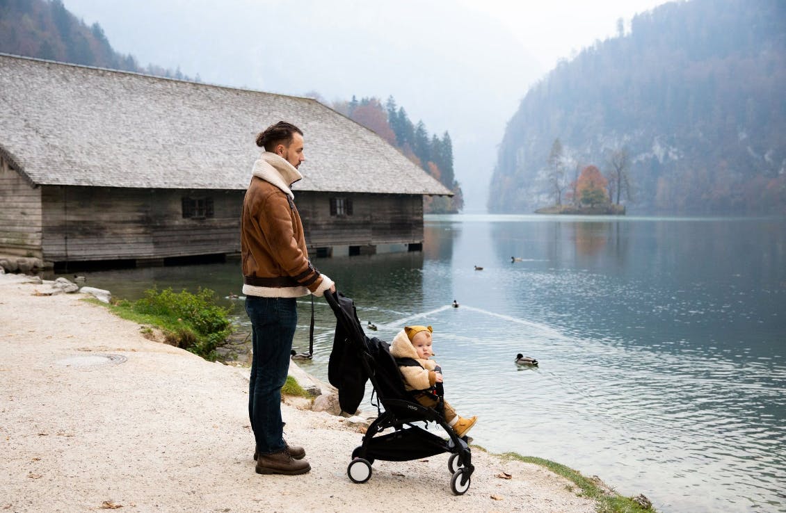 A man and a baby stand at the edge of a lake. The baby is in a stroller.