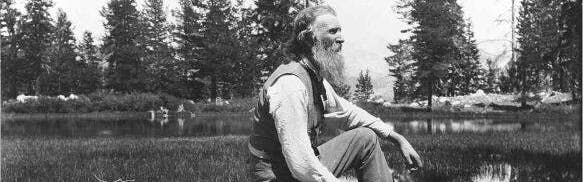 A black and white photo of John Muir sitting by a lake lined with trees