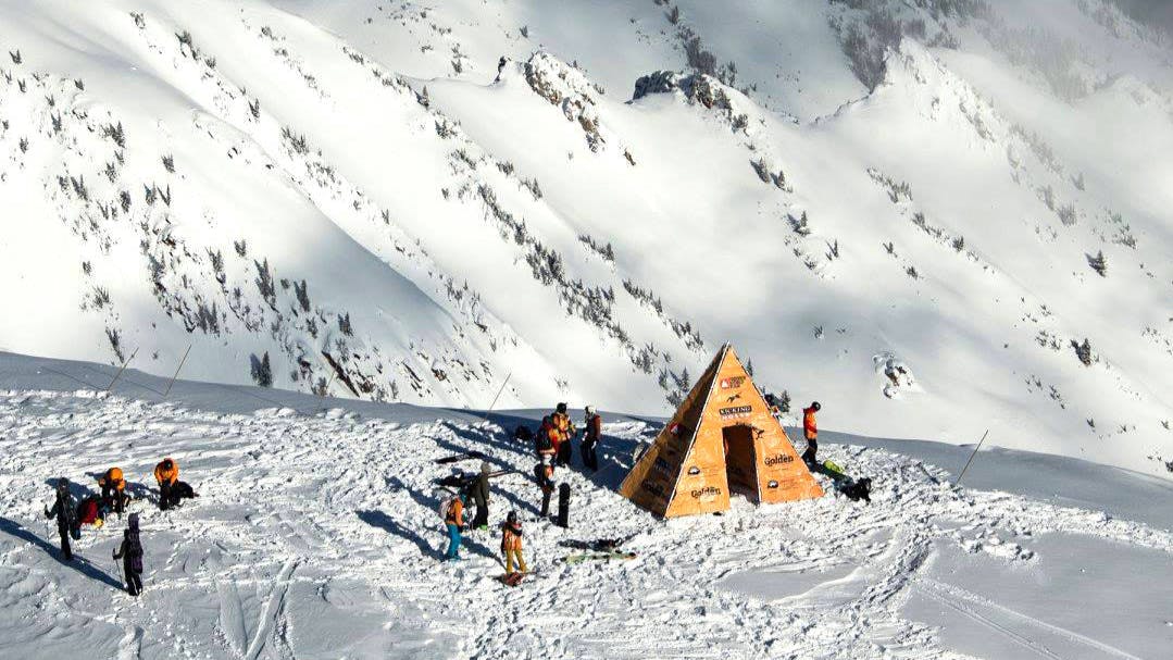 A triangular hut is set up at the top of the mountain and athletes dressed in orange mill around it. 
