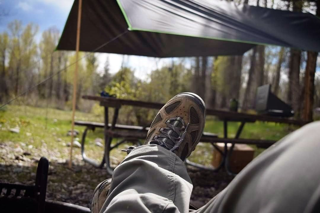 A view of a lounging hiker's grey pants and hiking boots, with a picnic table covered with a tent canopy in the background