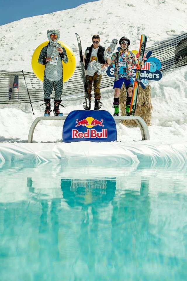 Three skiers on a podium holding their skis. There is a pond in front of them, a ski hill in the back, and the podium reads "Red Bull".