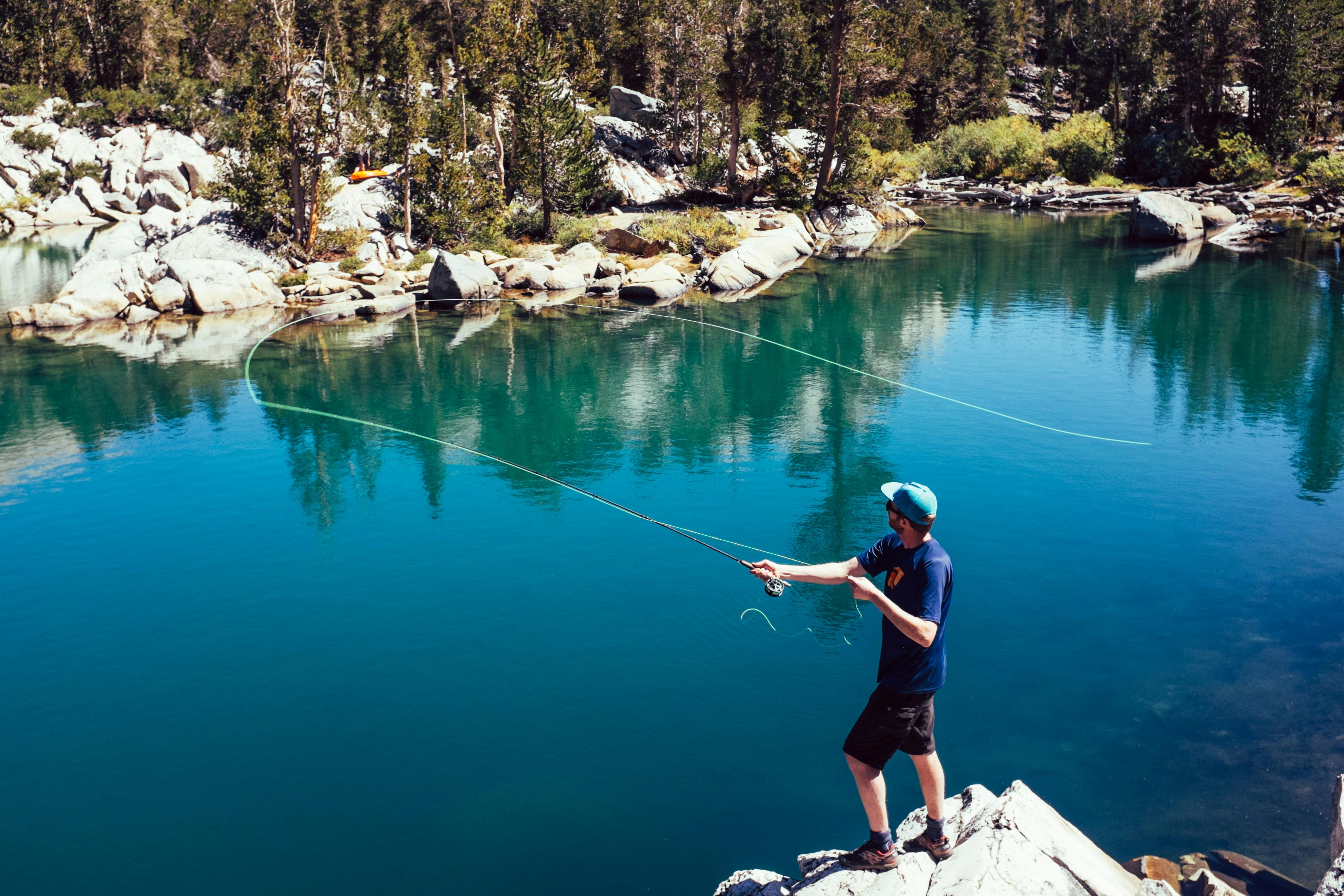 A man in shorts and a t shirt casts a fly fishing rod while standing on some rocks