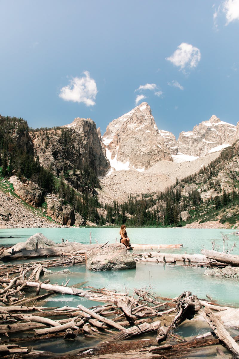 The author sits on a boulder that is surrounded by logs floating in a turquoise-colored alpine lake. The sky is baby blue above. 