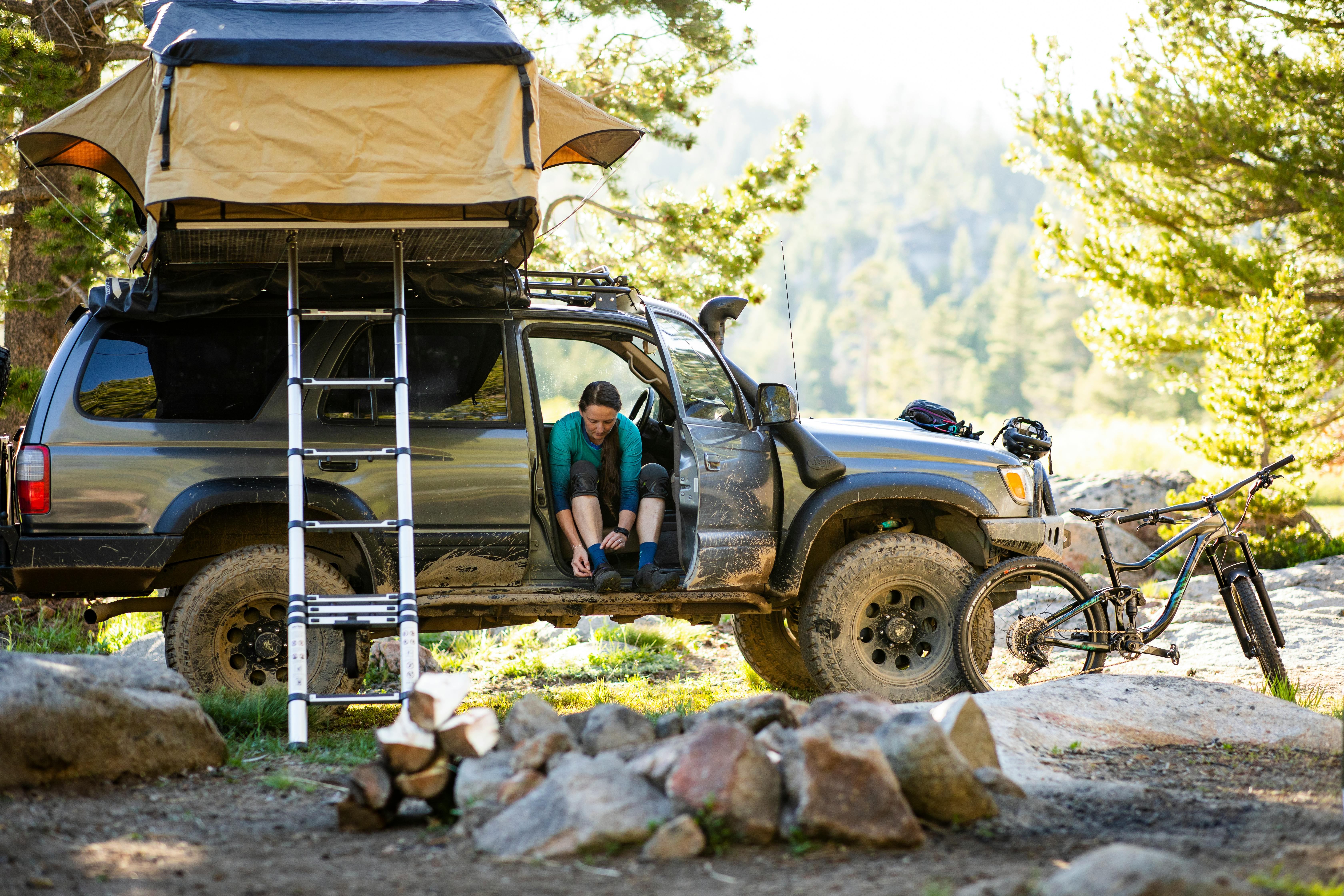 A woman sitting in her jeep putting on her shoes, a tent installed on the roof of the jeep and her mountain bike standing off to the side.