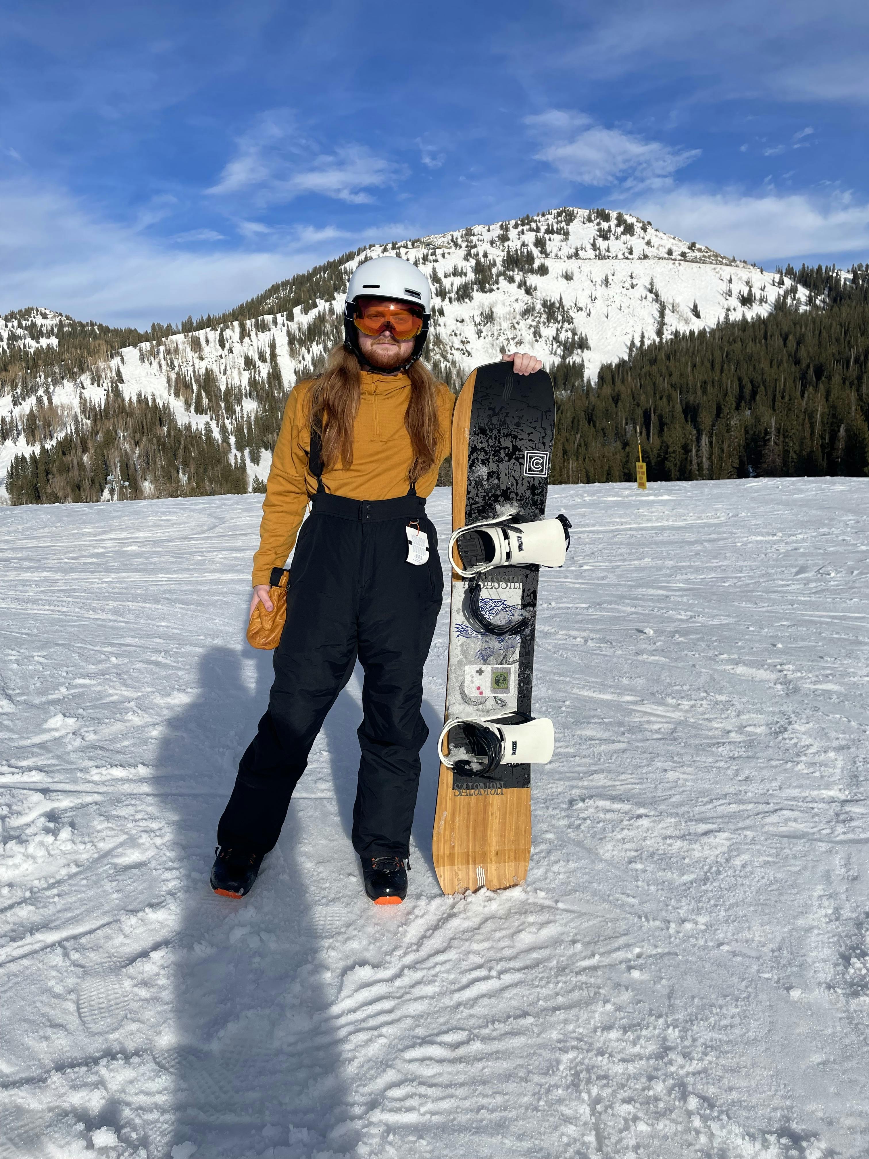 A man posing with the Salomon Assassin Snowboard · 2022. He is standing at a ski area and there are snowy mountains visible in the distance. 