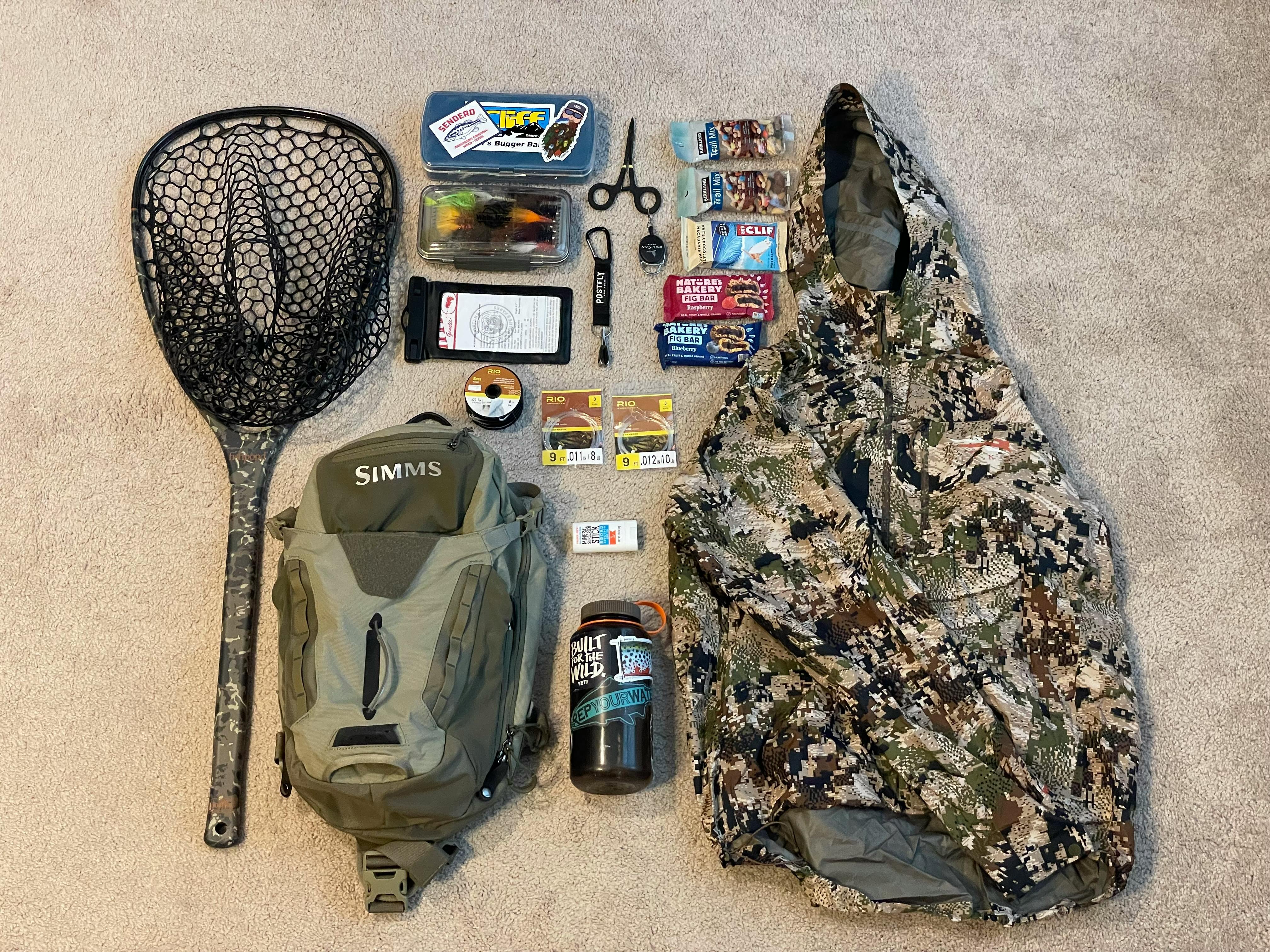 The author has laid out his pack, his net, a camp jacket, a water bottle, tools, tippet, snacks, and more on a carpet.