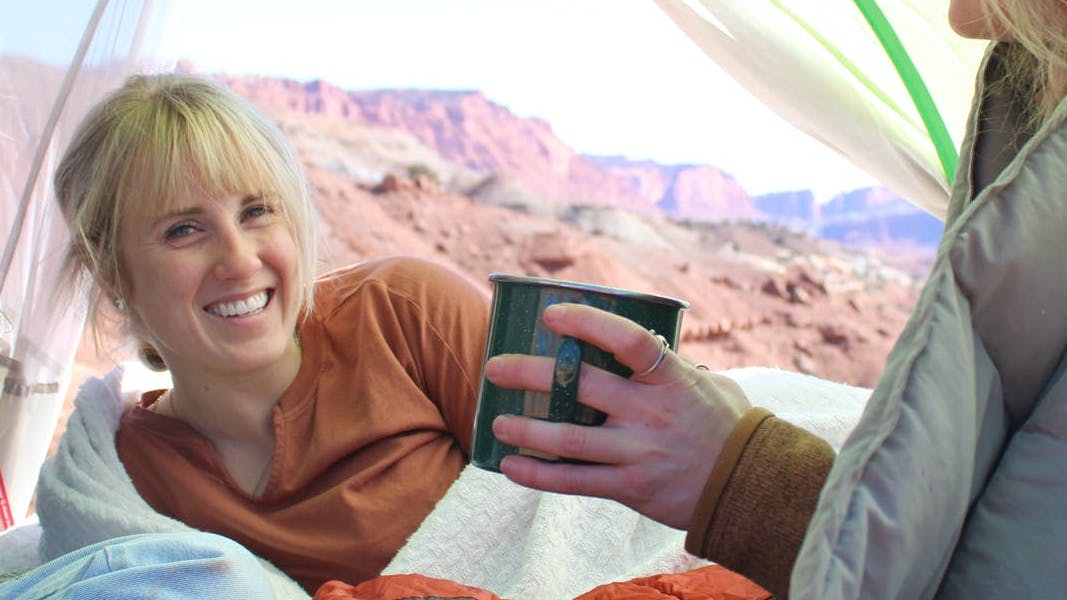 Three campers sit in a tent together laughing. One is looking at the camera and one is holding a mug. There are mountains in the background. 