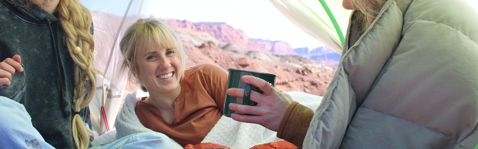 Three campers sit in a tent together laughing. One is looking at the camera and one is holding a mug. There are mountains in the background. 