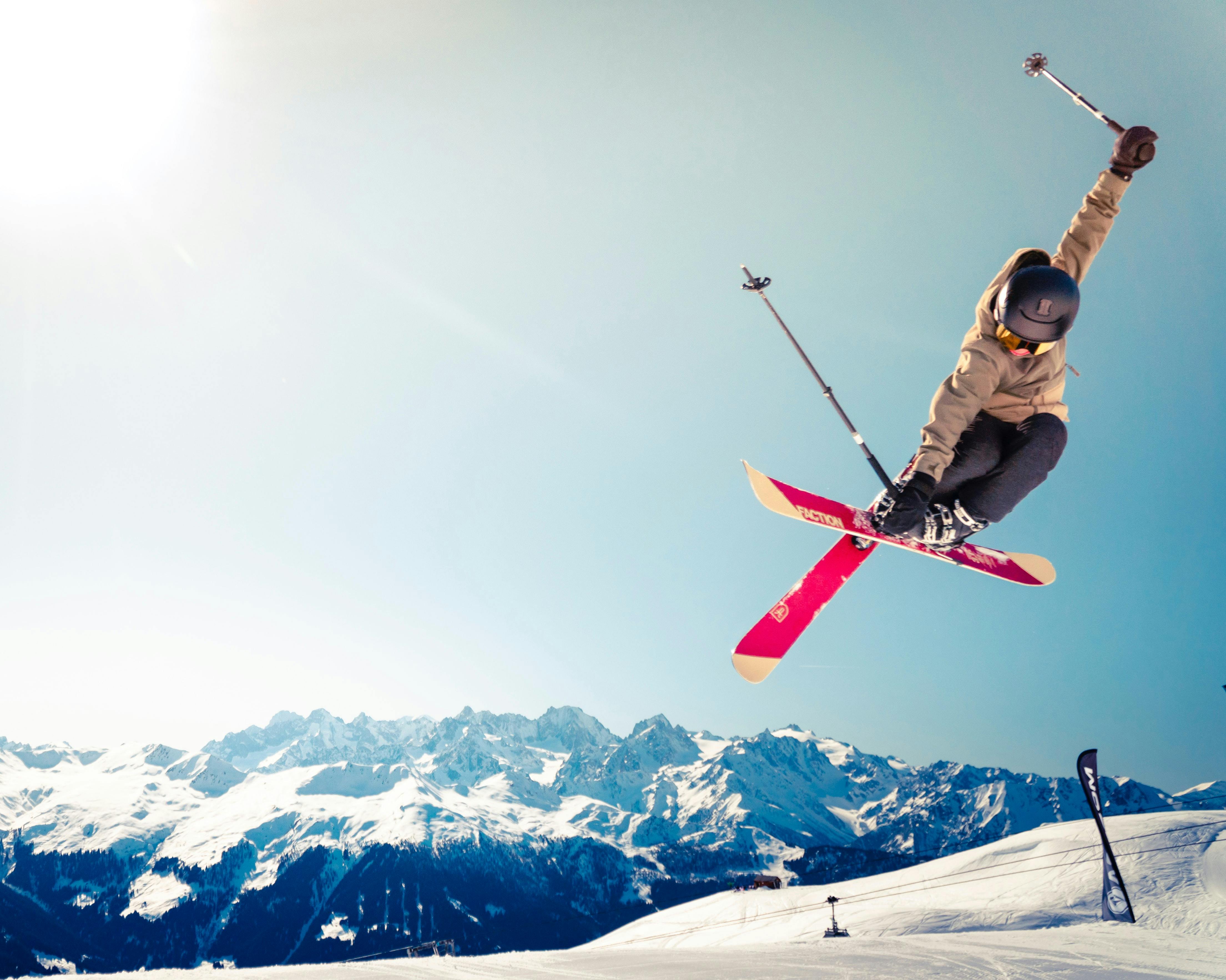 A skier flying through the air, arms outstretched and skis crossed in an epic trick - the sun blazing down on the mountains in the background.