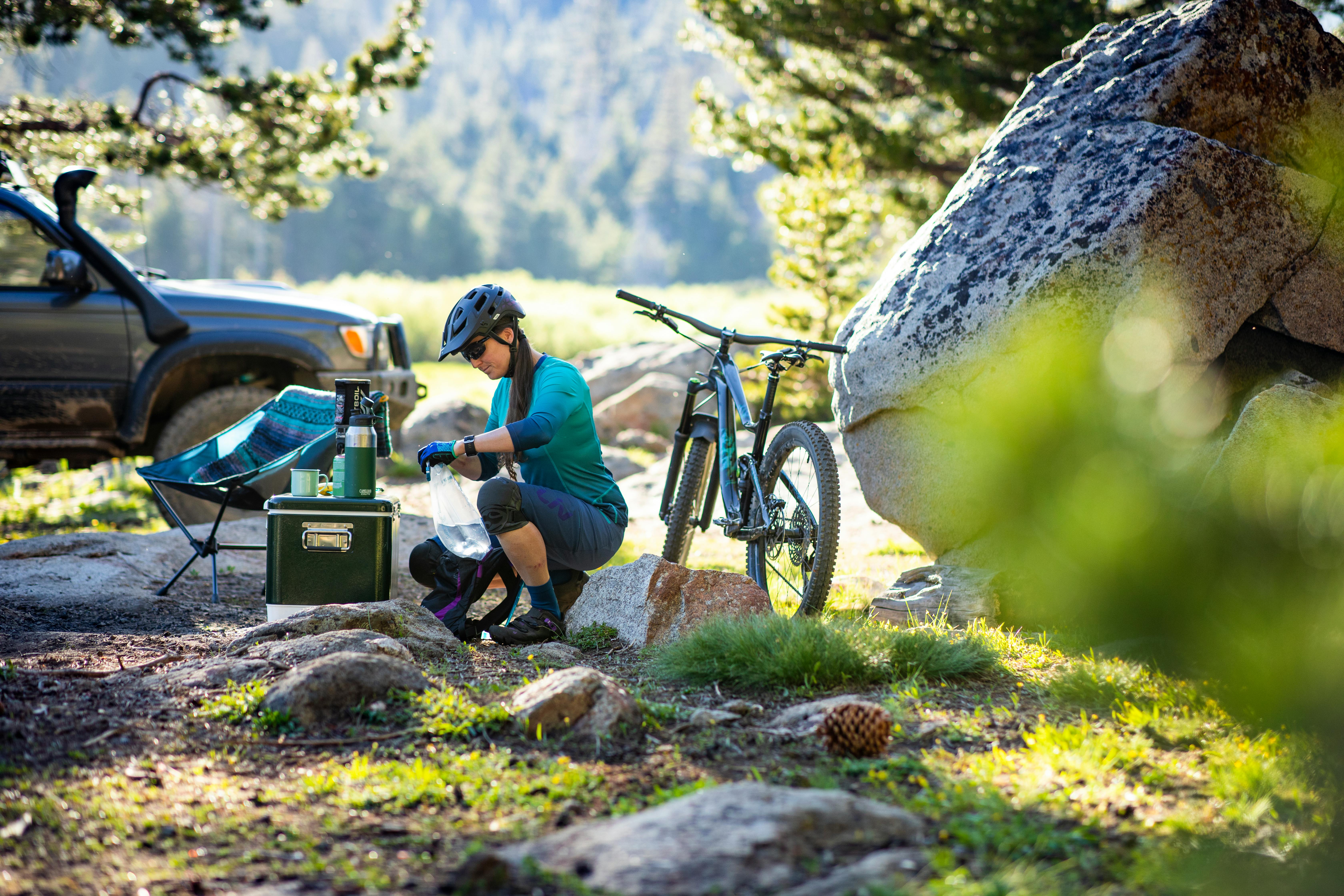 A woman at a campsite with her water bottle, cooler, camping thermos, mugs, and camping chair, and with her mountain bike behind her.
