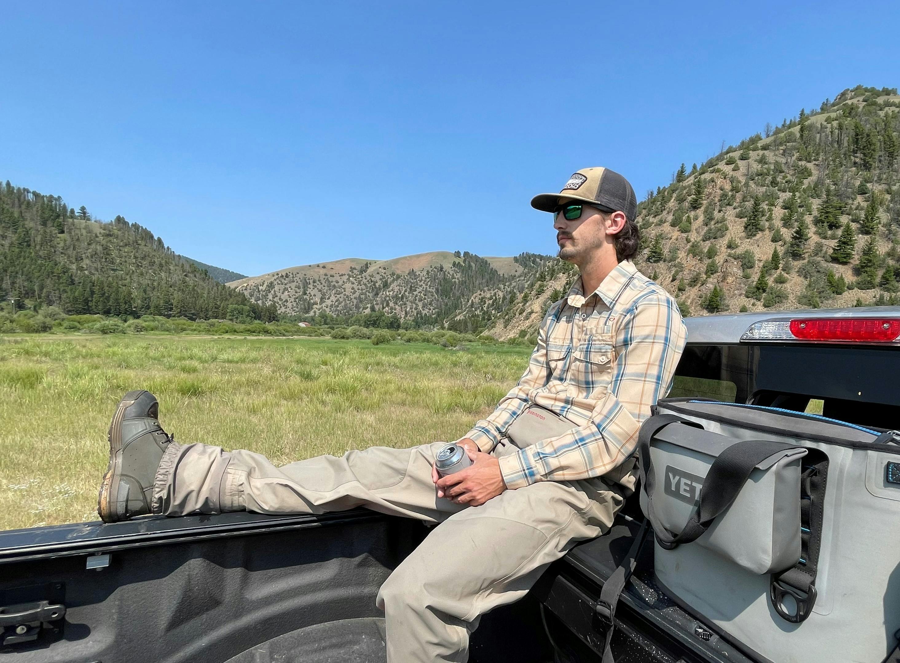 The author sits in the back of his truck, next to a Yeti cooler. He wears waders, boots, and drinks a beer looking out into the distance.