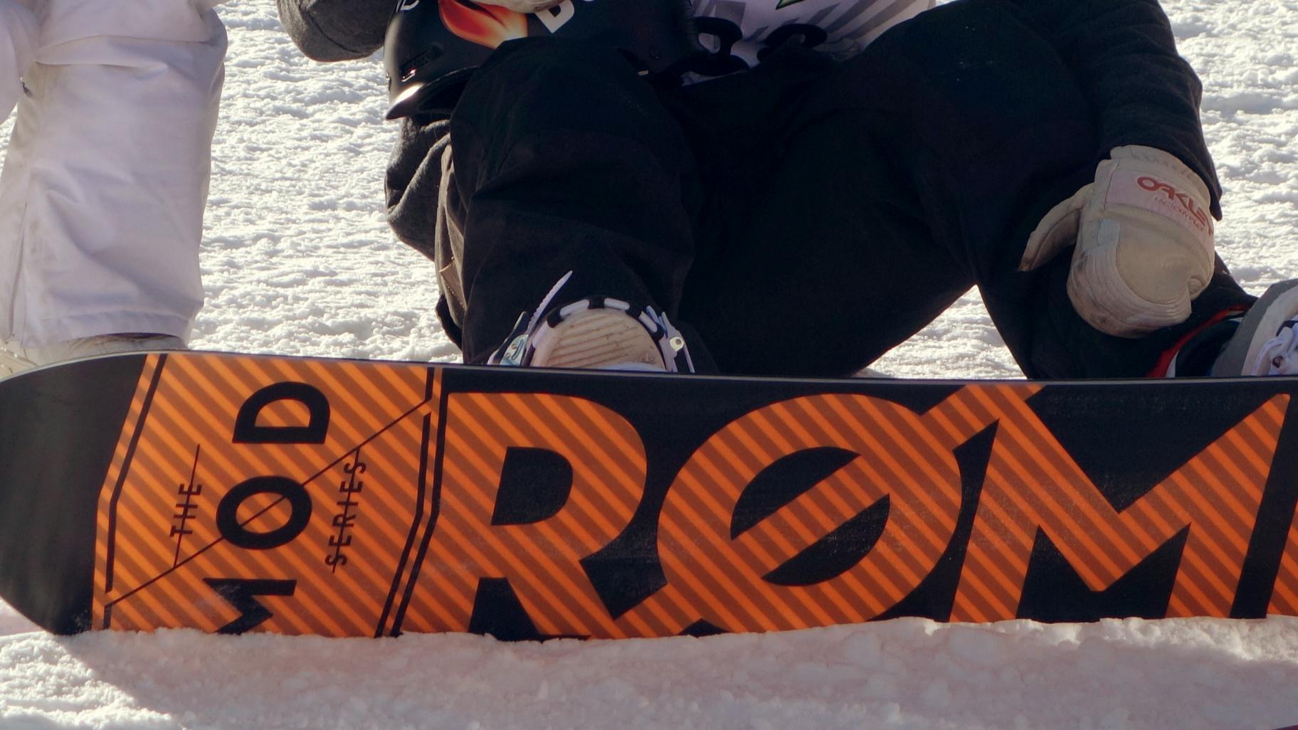 A snowboarder sitting on the ground with the bottom of his snowboard visible. The bottom of his snowboard says "ROME the MOD Series".