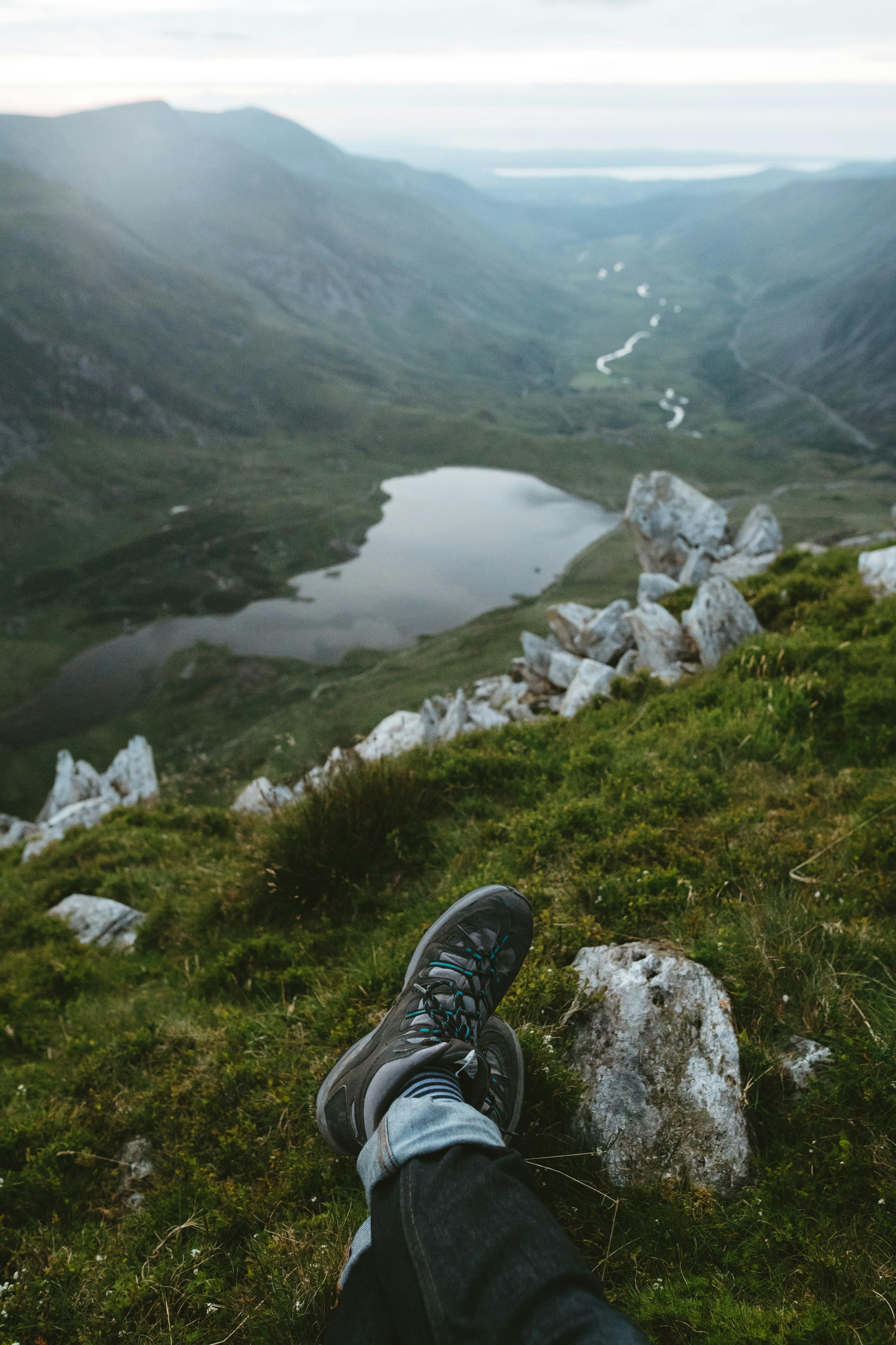 The photo is focused on someone's outstretched legs and their hiking boots. They sit on a rocky hillside with an alpine lake and river stretching into the background. 