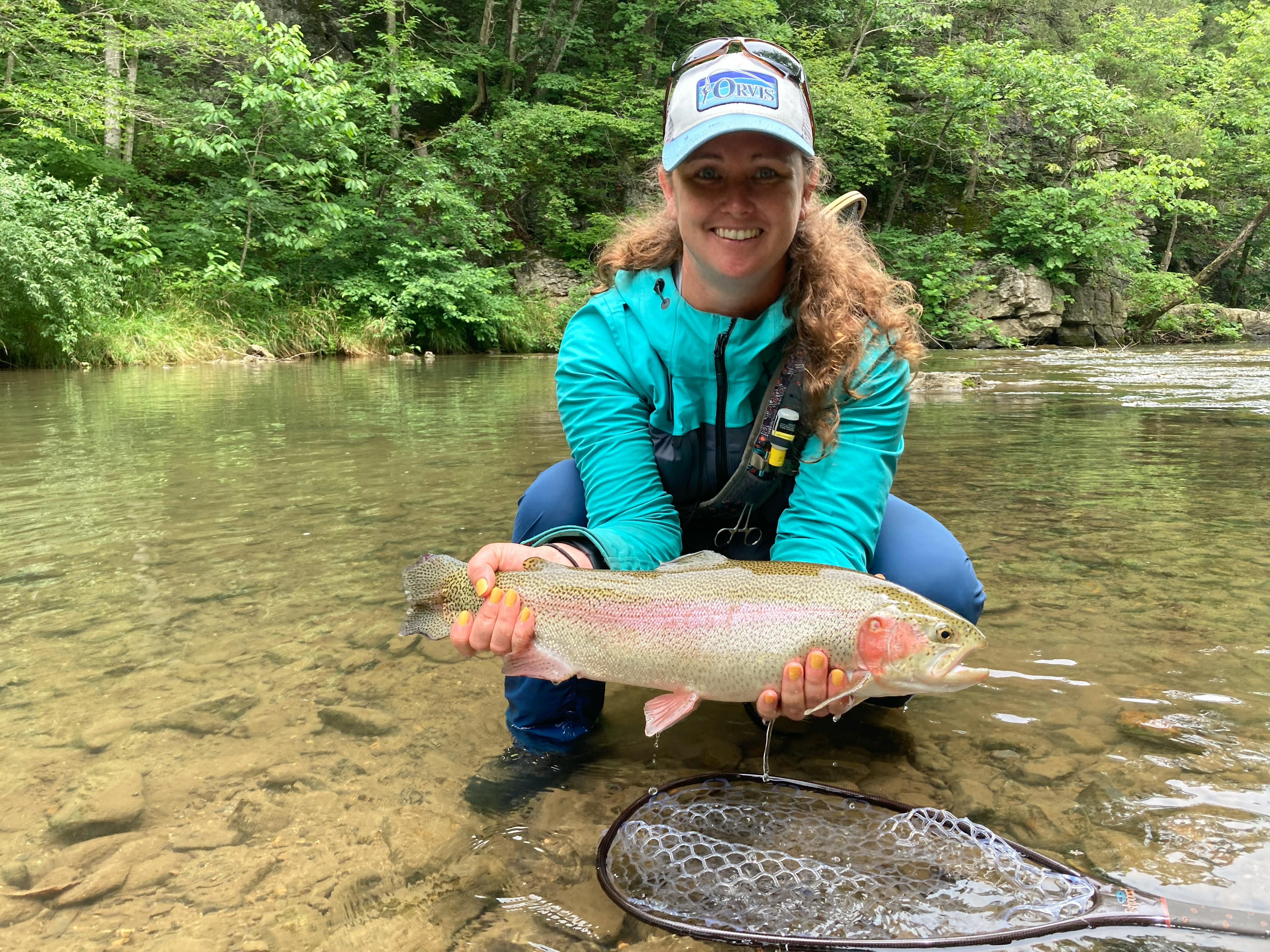 The author squats above a creek in waders while holding a large brown trout out of the water above her floating net. She has a big smile on her face.