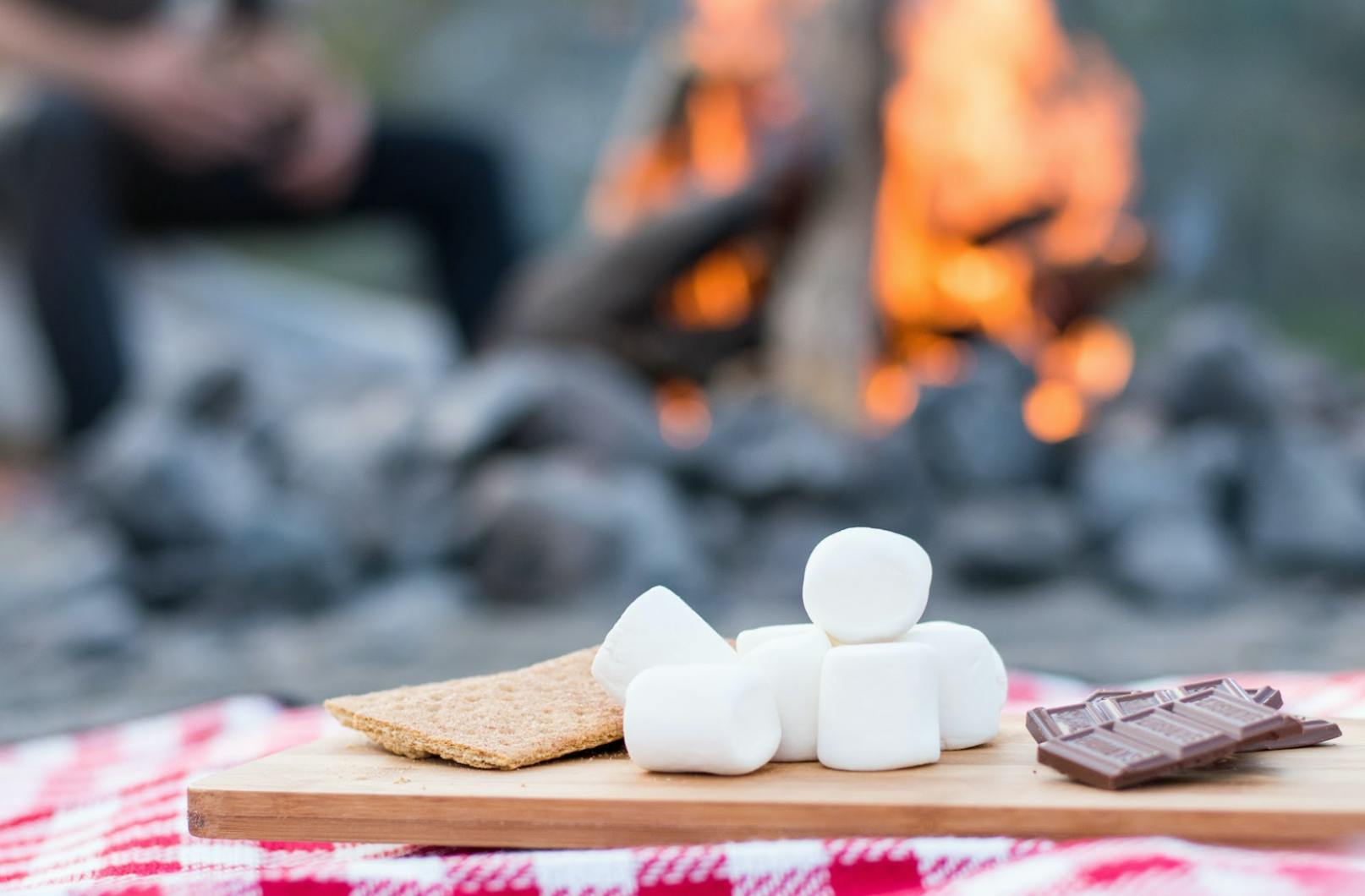 A graham cracker, chocolate, and marshmallows on a table with a fire in the background.