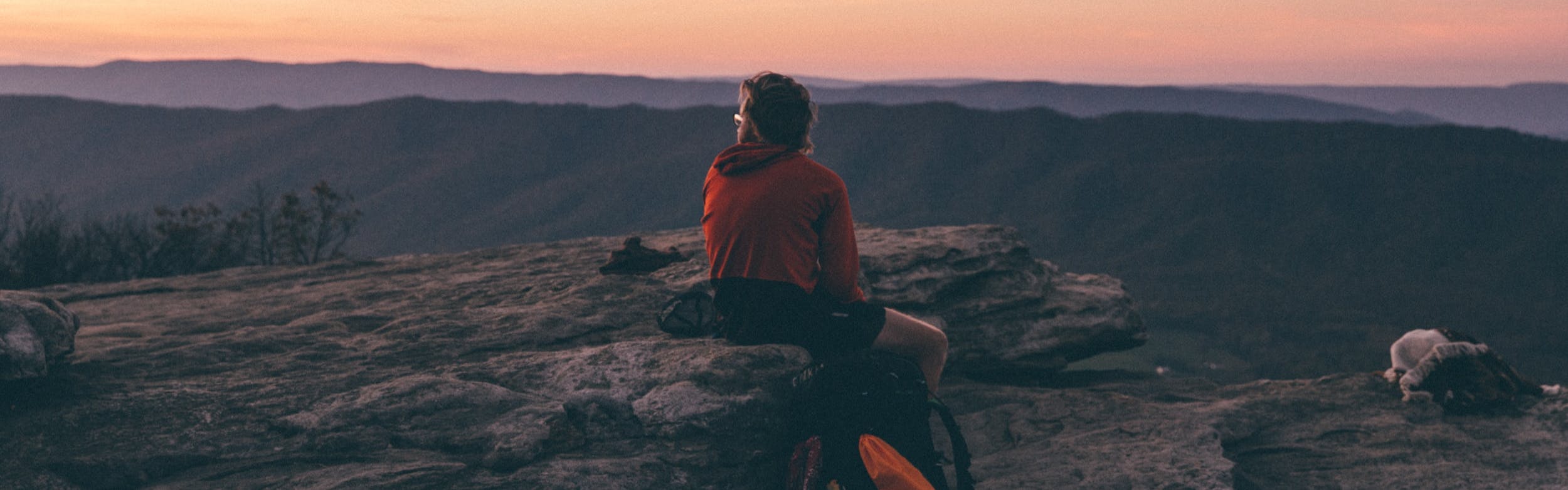 A man sits on a rock overlooking mountains on a sunset.