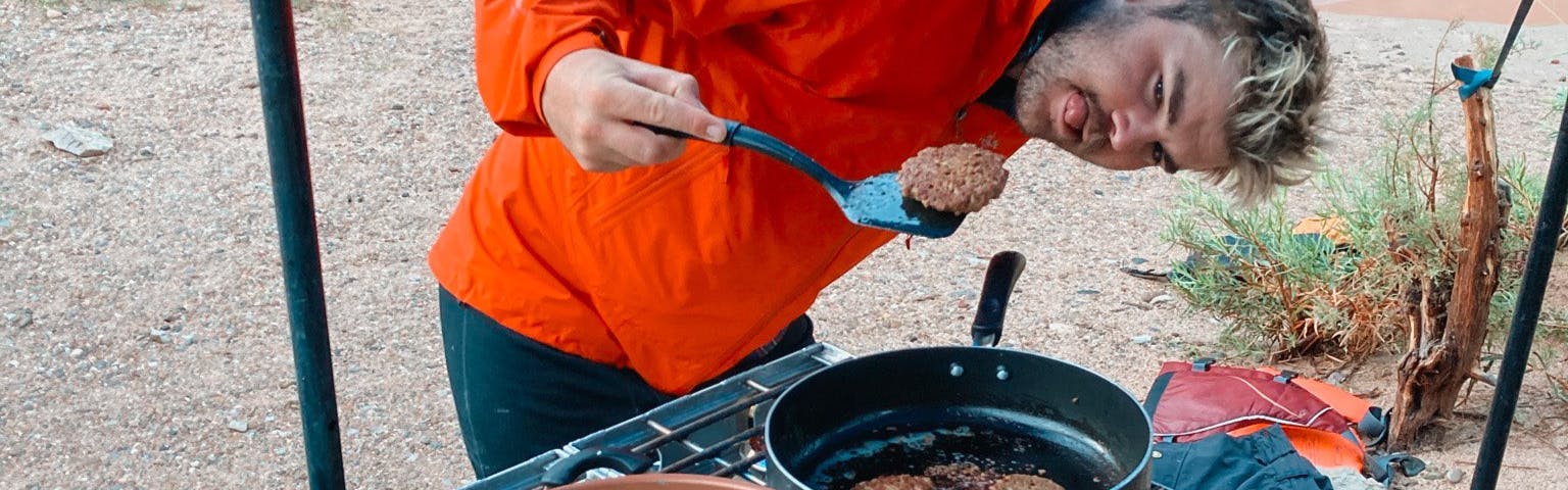 A camper making food and looking at a veggie sausage that he's holding on a spatula. 