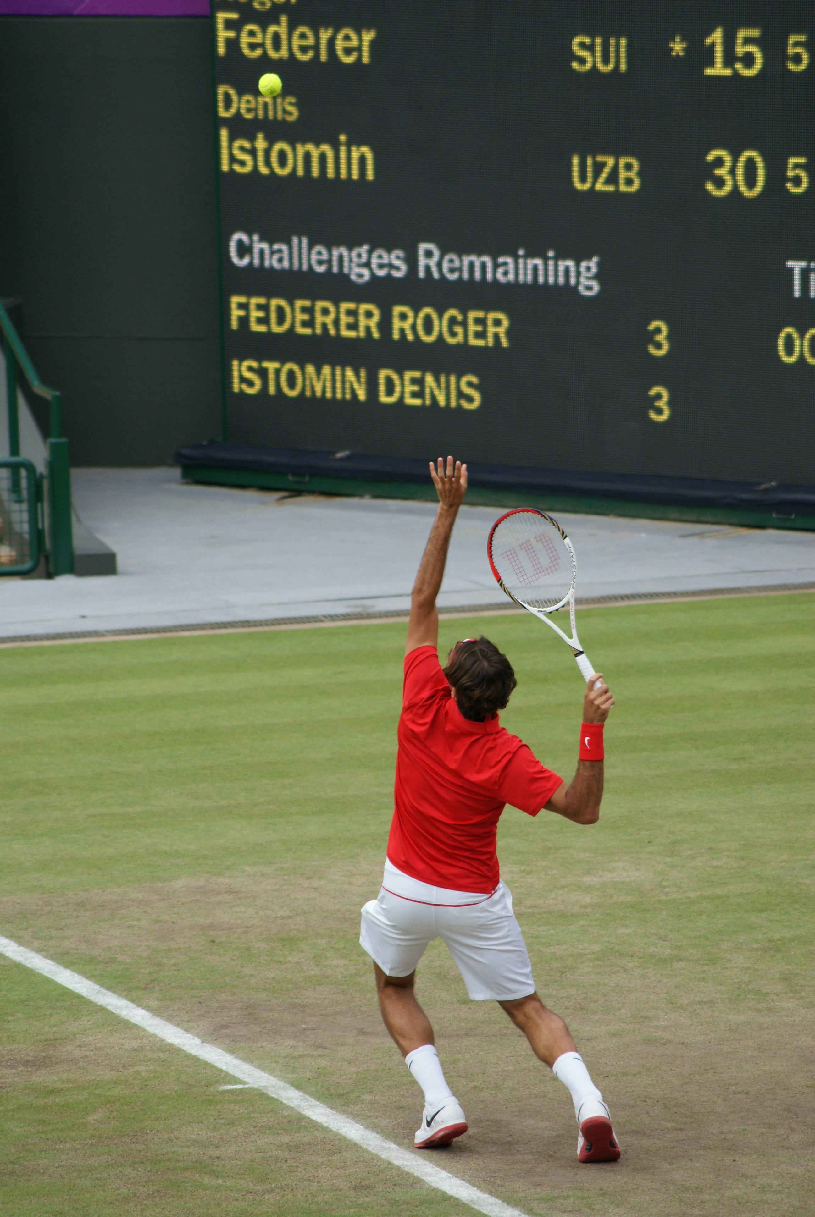 A tennis player in a red shirt and white shorts about to hit a tennis ball flying through the air with an overhead swing