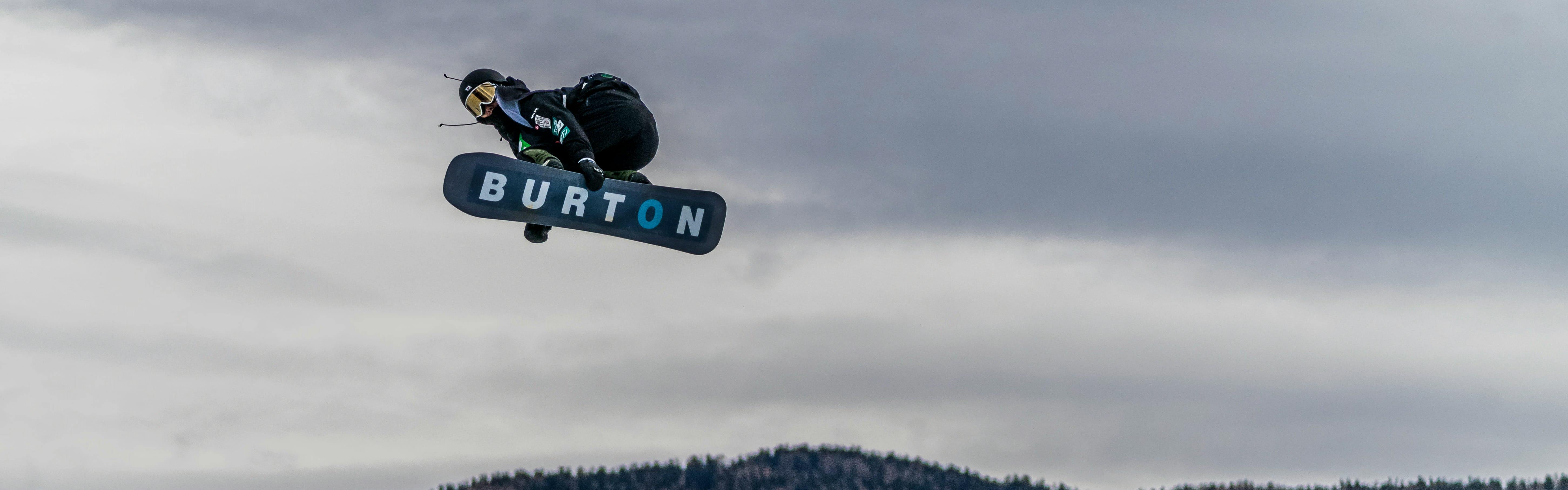 A snowboarder jumping in the air and grabbing his snowboard. The bottom of the snowboard reads "Burton". 