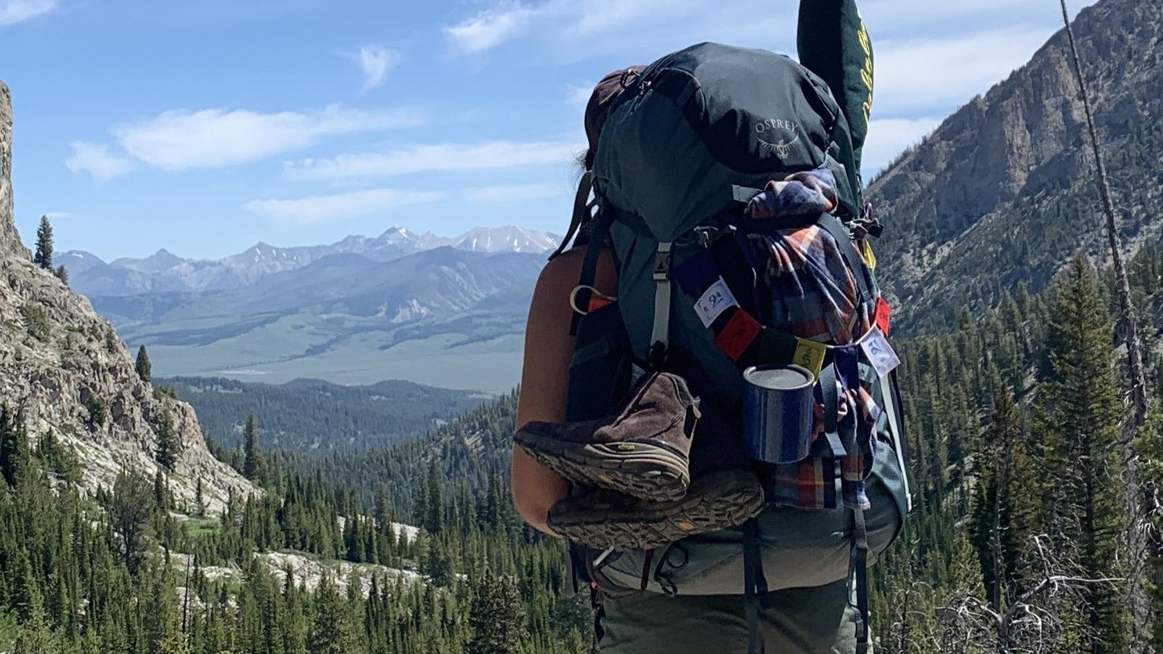 A woman with the Osprey Ariel 75 L backpacking backpack stands at the top of a lookout. There are mountains and rocky cliffs in the distance and attached to her pack are some shoes, a cup, and other camping equiptment.