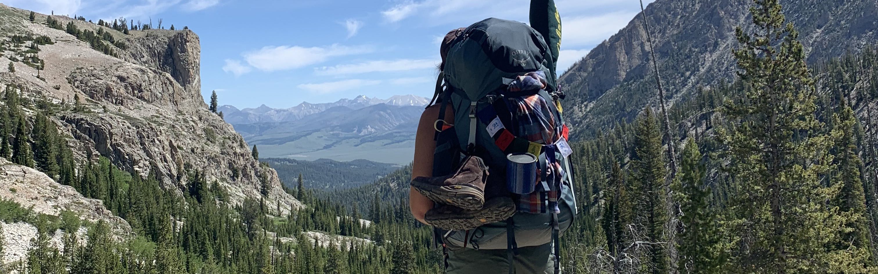 A woman with the Osprey Ariel 75 L backpacking backpack stands at the top of a lookout. There are mountains and rocky cliffs in the distance and attached to her pack are some shoes, a cup, and other camping equiptment.
