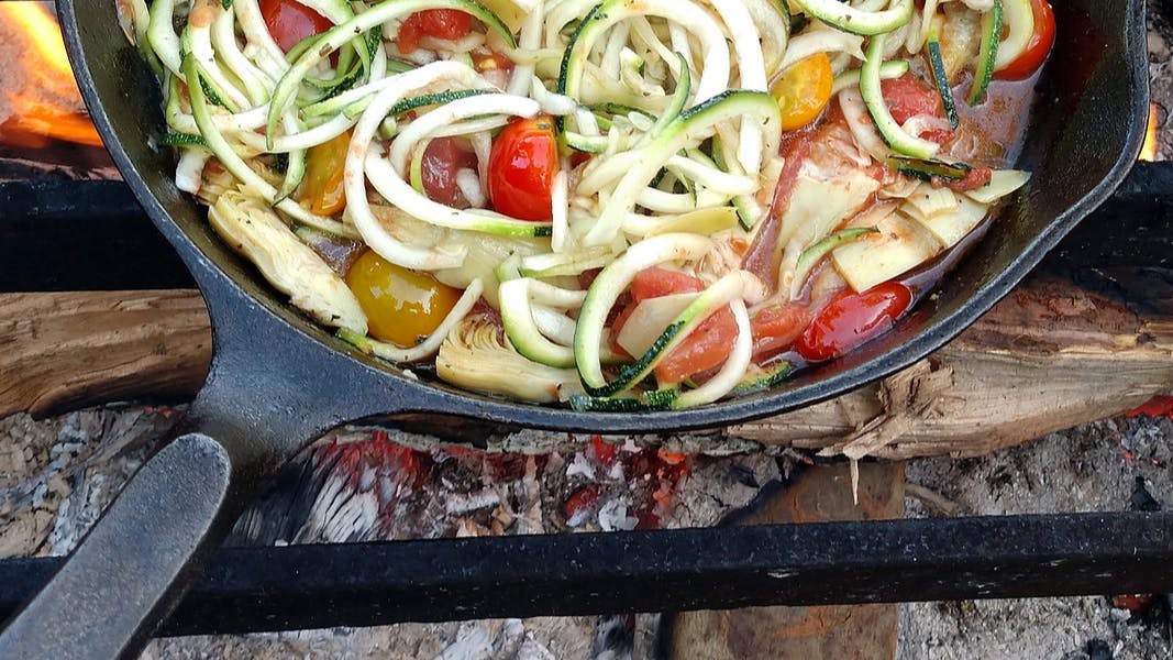A cast-iron skillet full of chopped vegetable sautees on a metal grate over a campfire.