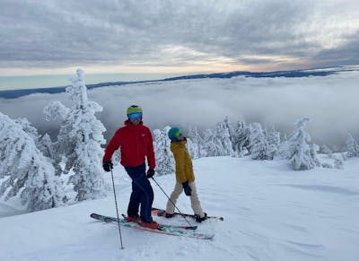 Two people stand on a very snowy trail. One is a skier and one is a snowboarder. There are snowy trees in the background and clouds in the distance.