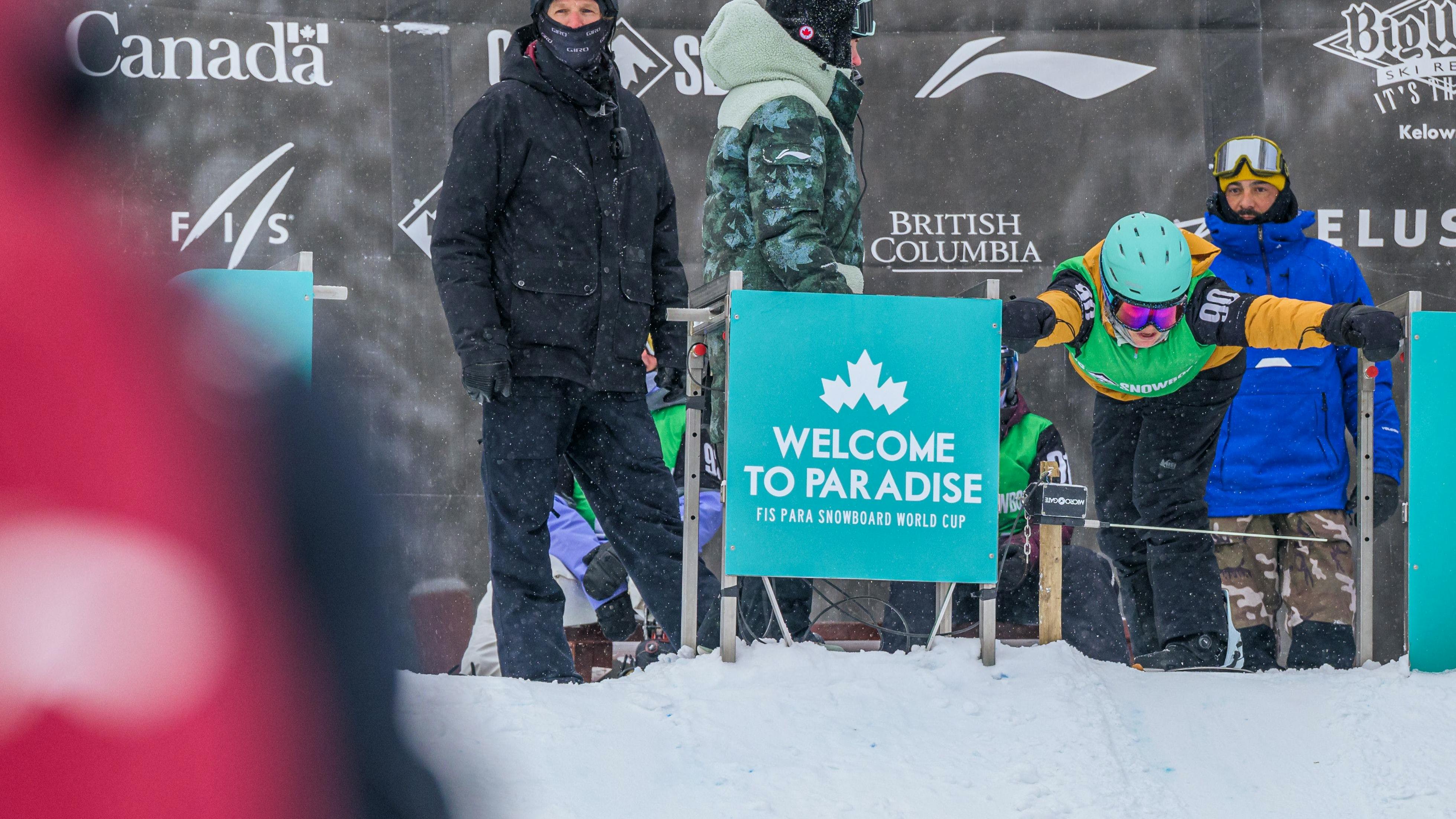 Snowboarder in yellow jacket, green race bib and blue helmet bent over in the starting gate at the start of a banked slalome race with other riders standing in the background.