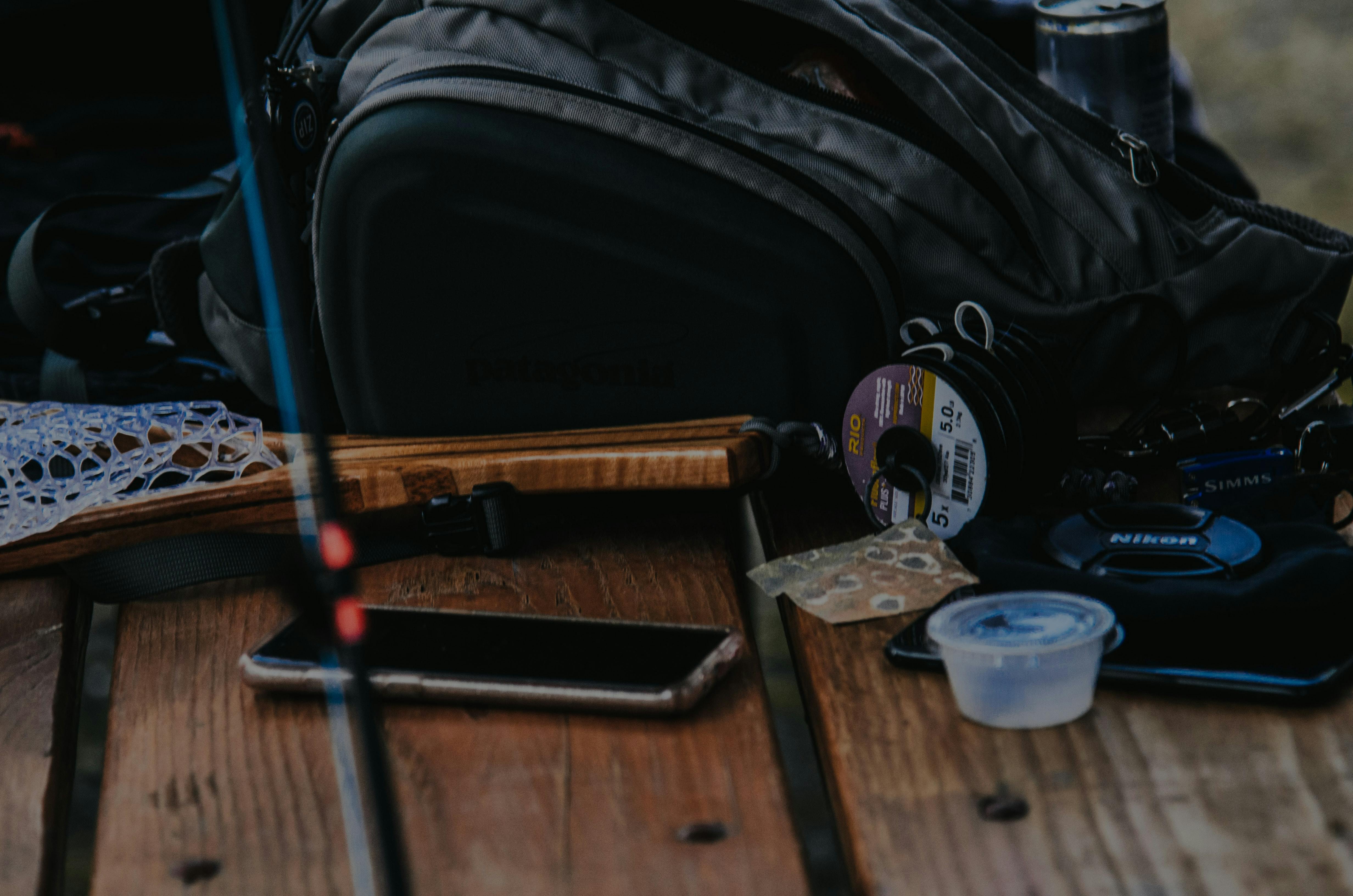 Fishing gear, including a net, rod, tippet, and a phone, sitting on a wooden picnic table. 