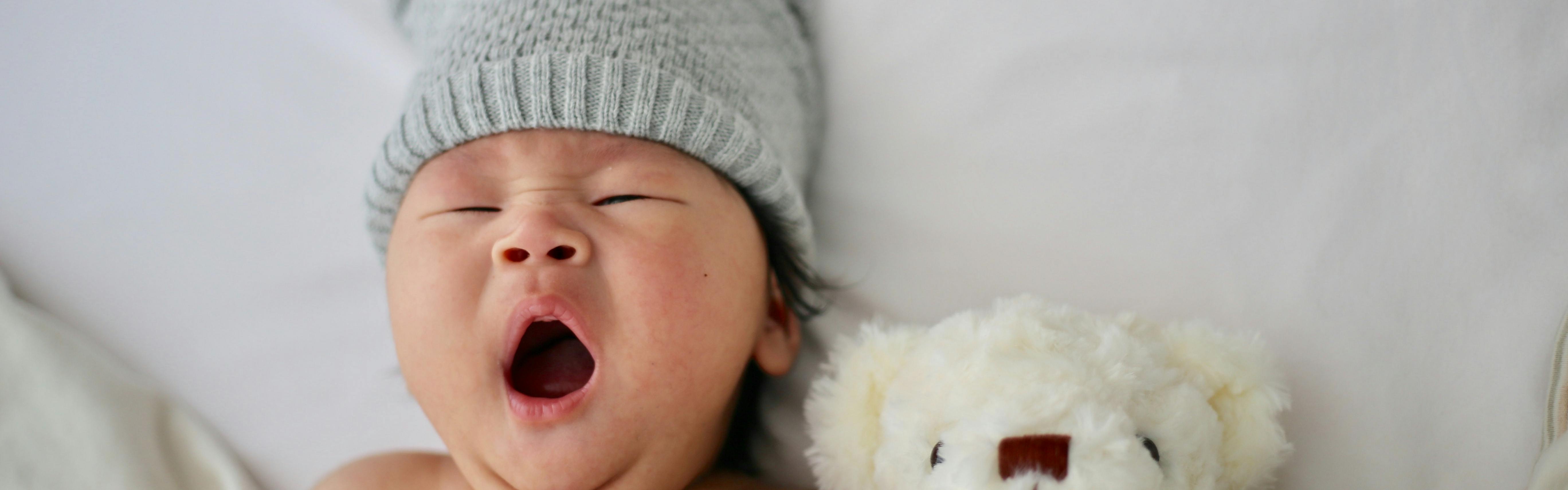 A baby wearing a hat takes a big yawn. His teddy bear is next to him.