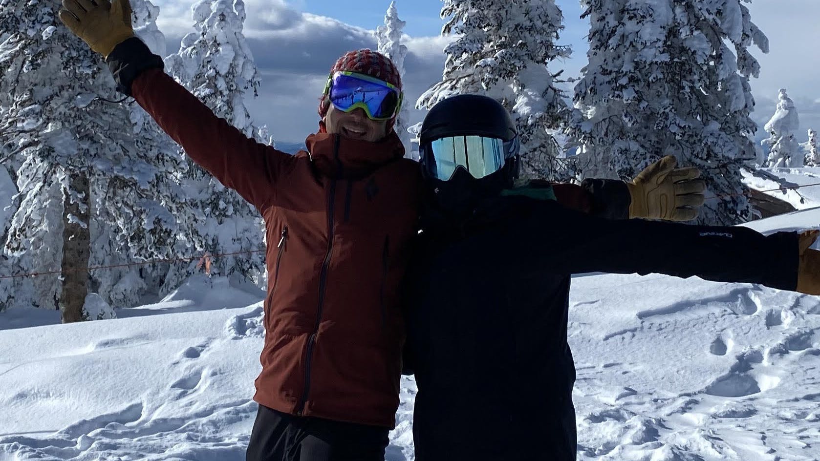 Two skiers stand at the top of a snowy mountain with their arms outstretched. There are snowy trees behind them. 