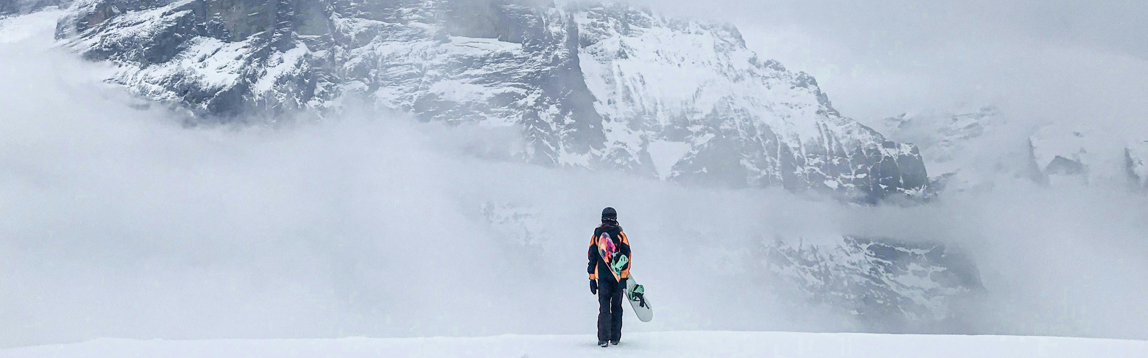 A snowboarder stands with their back to the camera, looking at mountains