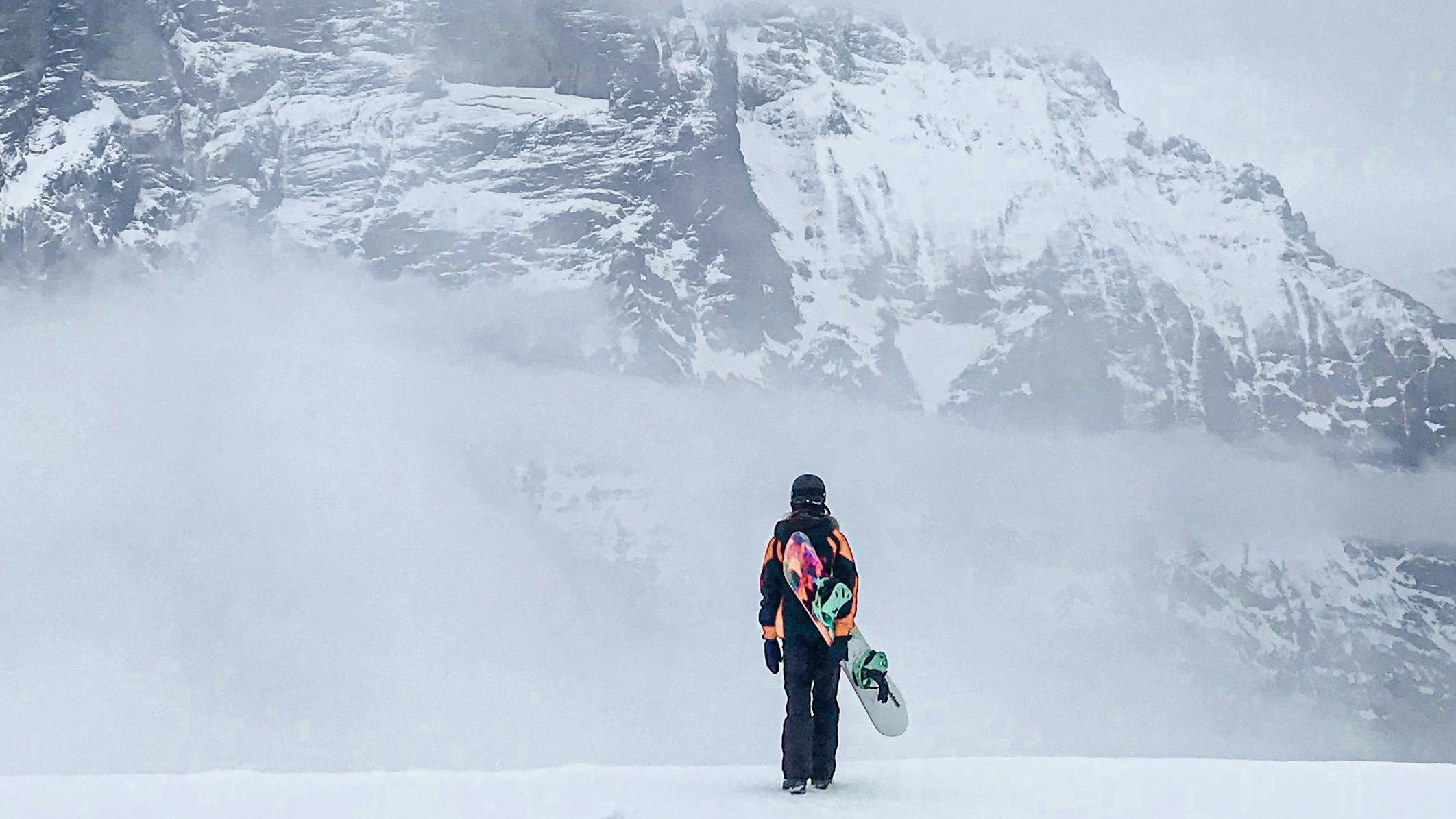 A snowboarder stands with their back to the camera, looking at mountains