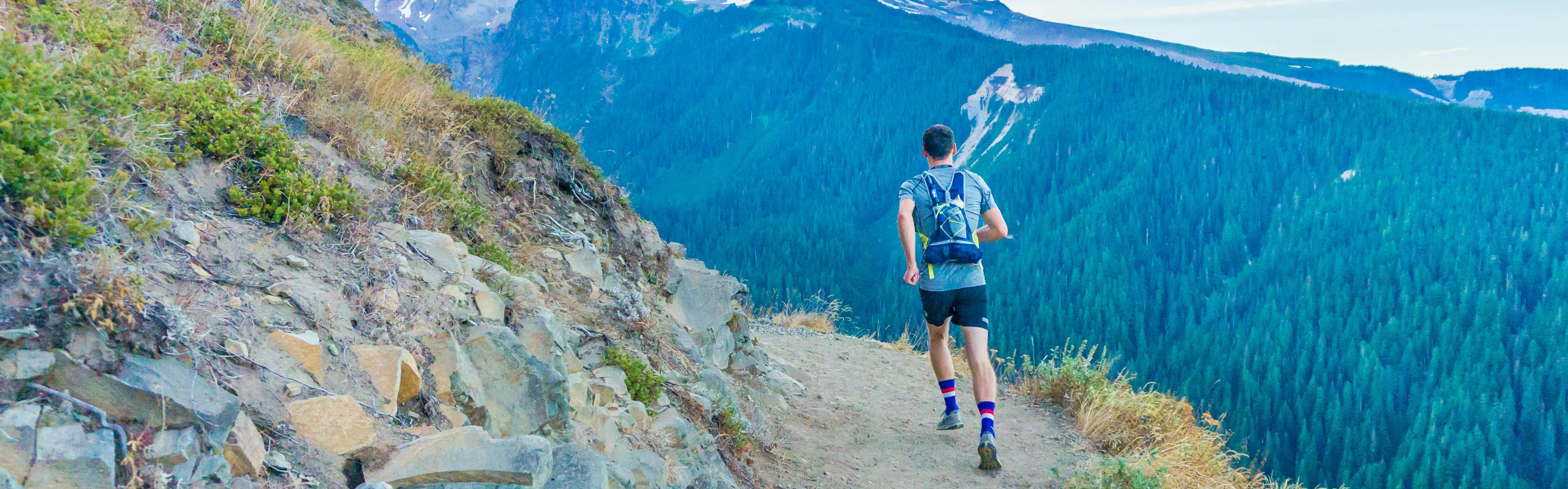 A man with a running vest jogs along a mountainside trail in trail runners. 