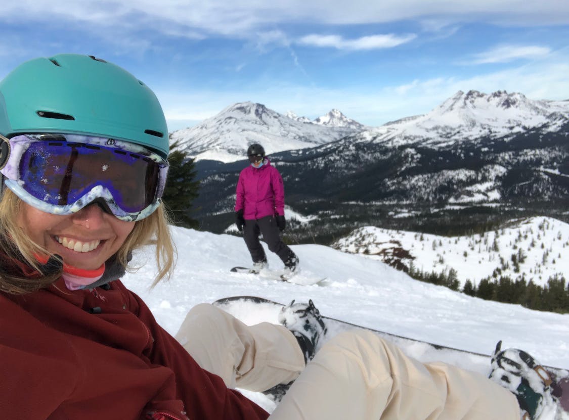 Two women are snowboarding. There are snowy mountains behind them.