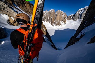 A man wearing the POC OBEX MIPS helmet is staring down at a couloir. He has his skis attached to his backpack in an A frame shape.