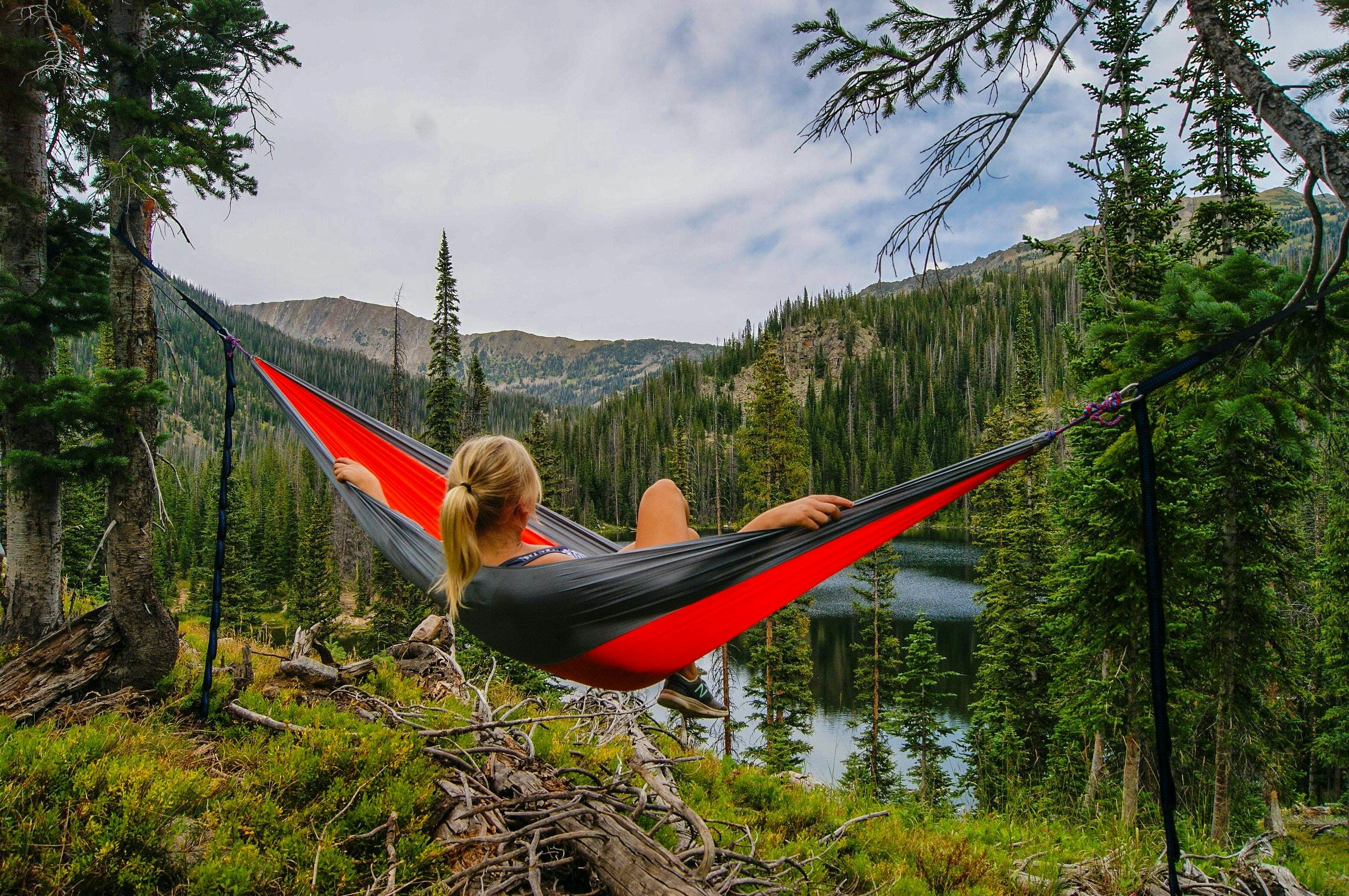 A woman leans back in a red and black hammock, looking out at trees and a lake